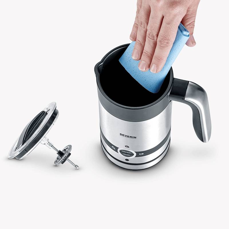 A hand is cleaning the inside of an electric milk frother with a blue sponge. The lid is lying beside it.