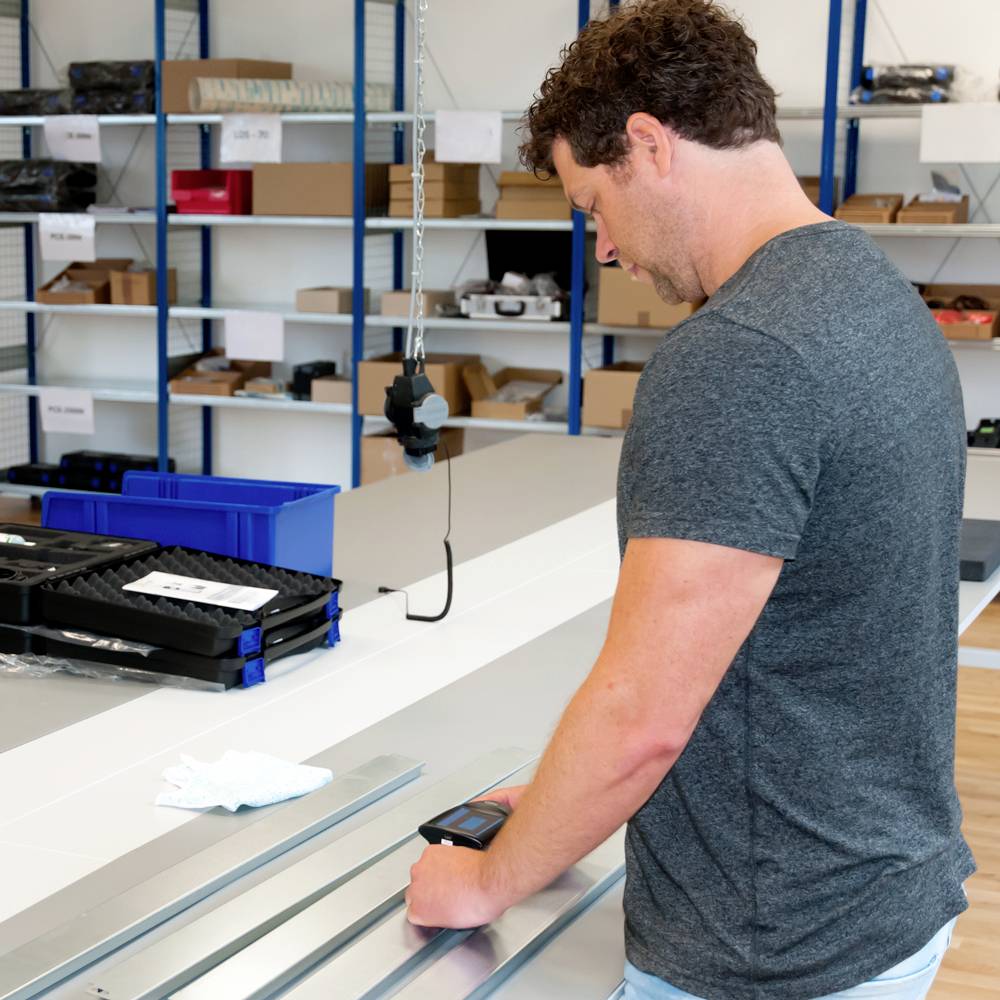 A man in a warehouse is using a handheld device to scan elongated metal parts, likely for inventory or quality control purposes.
