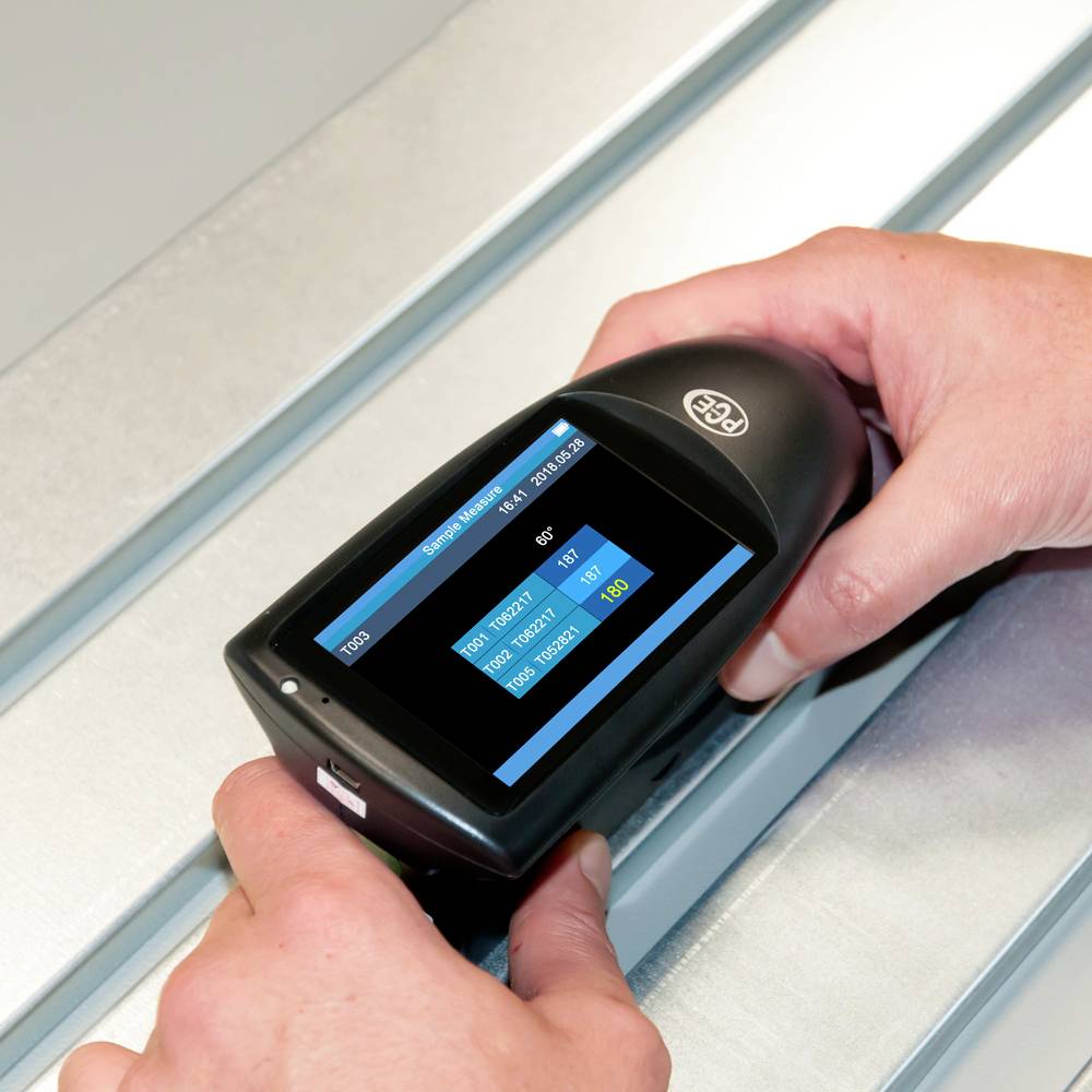 A person is holding a portable measuring device on a metal surface. The screen displays measurement values, including 'ISO' and '103'.