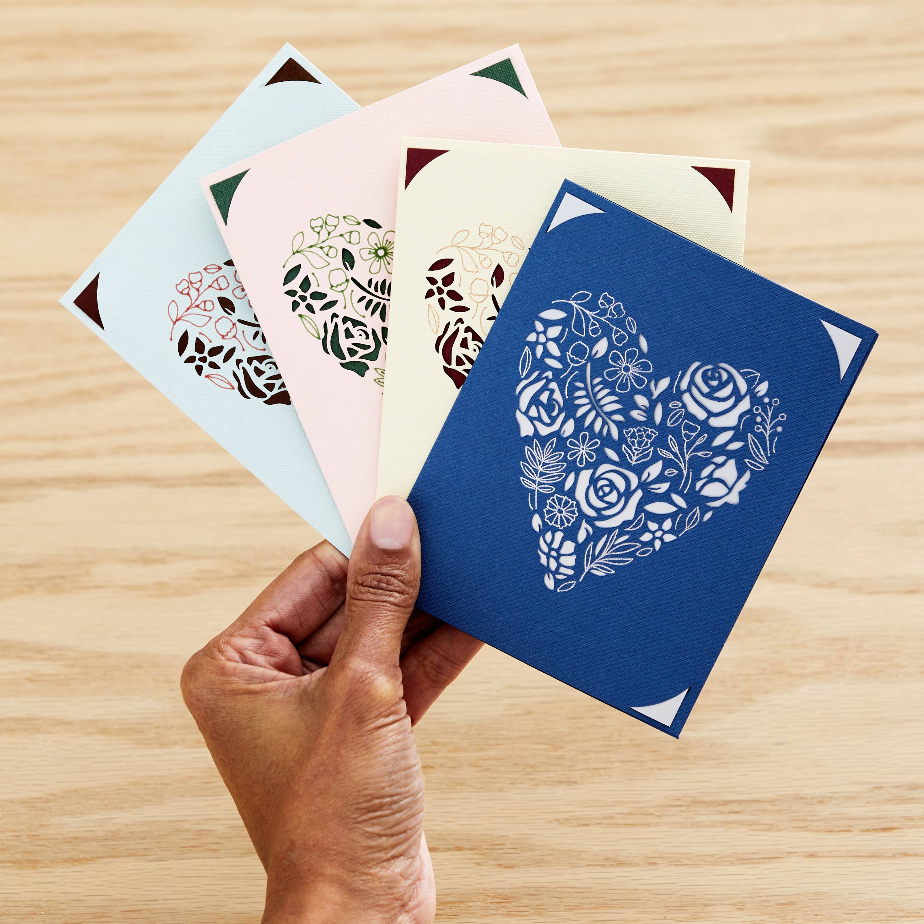 A hand is holding four greeting cards with laser-cut heart designs on a wooden table. Each card has a different colour.