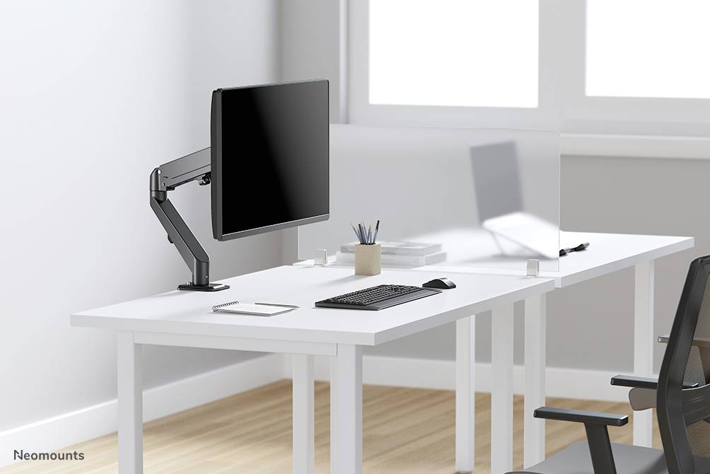 A modern, white desk with a monitor on an adjustable arm, a keyboard, and office supplies. Bright room with large windows.