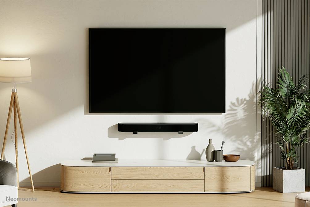 Modern living room interior with wall-mounted television, a soundbar beneath it and a wooden cabinet. A lamp on the left and a plant on the right.