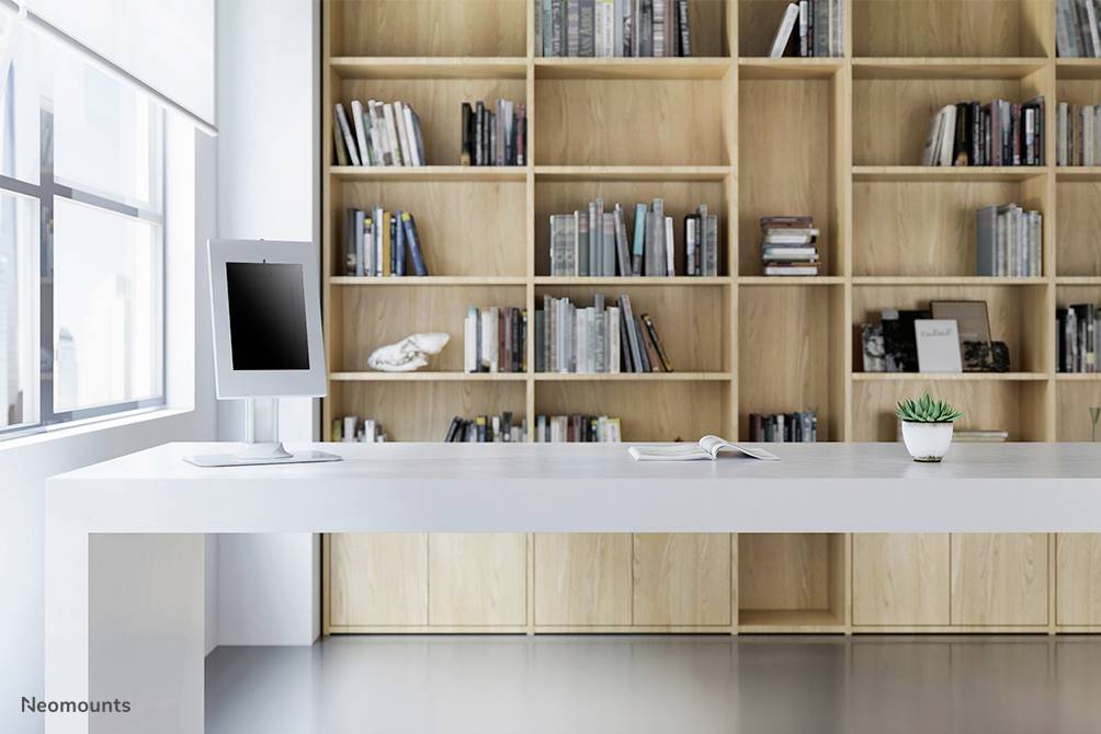 A minimalist workspace with a modern monitor on a white table, positioned in front of a large bookshelf, with a small plant beside it.