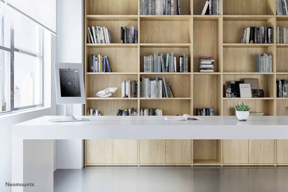 A modern, minimalist room with a white table, on which a monitor and a book are placed, in front of a wooden bookshelf filled with books.