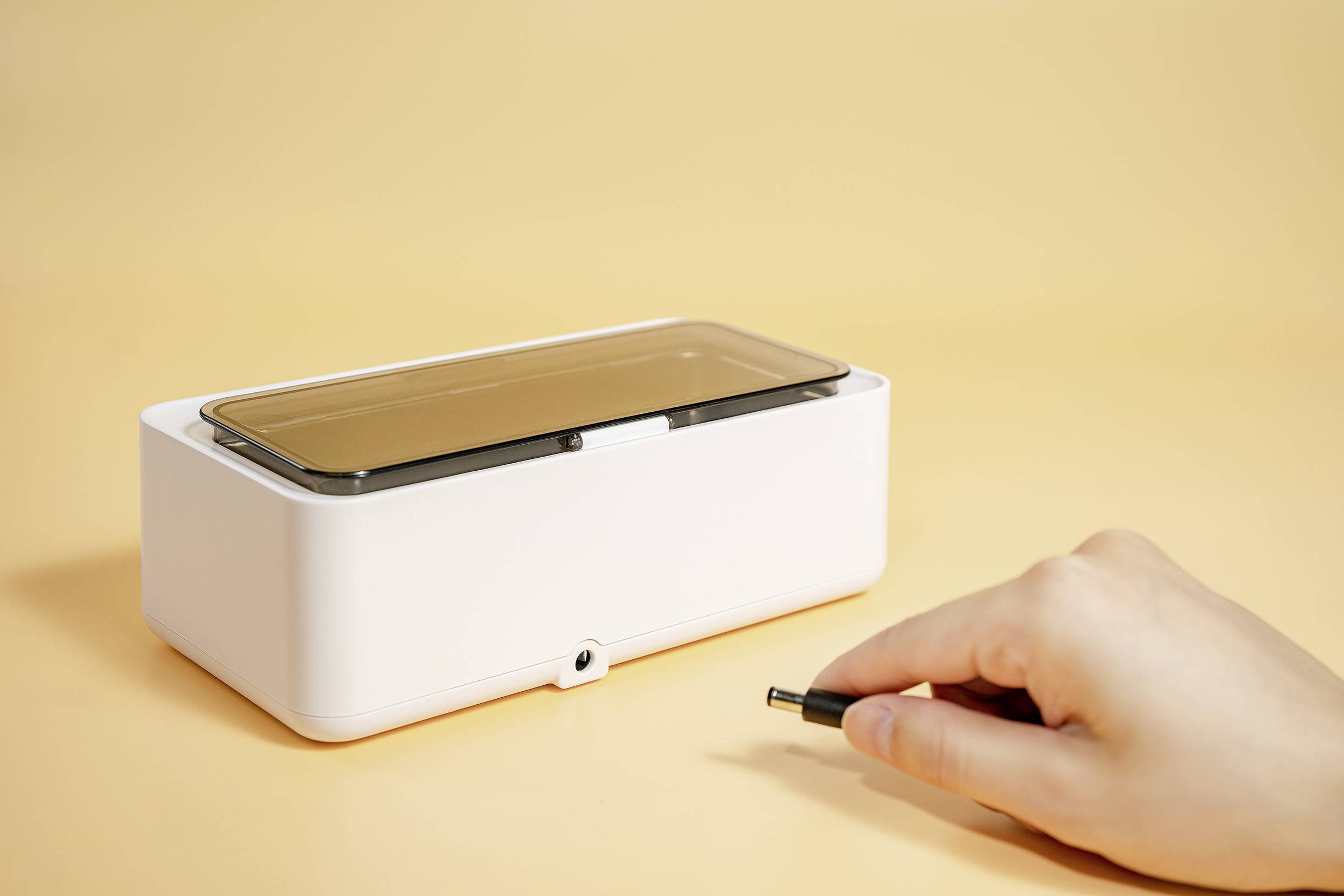 White UV disinfection box with lid, hand holding a power cable in front of it, on a beige background. Device used for sterilisation.