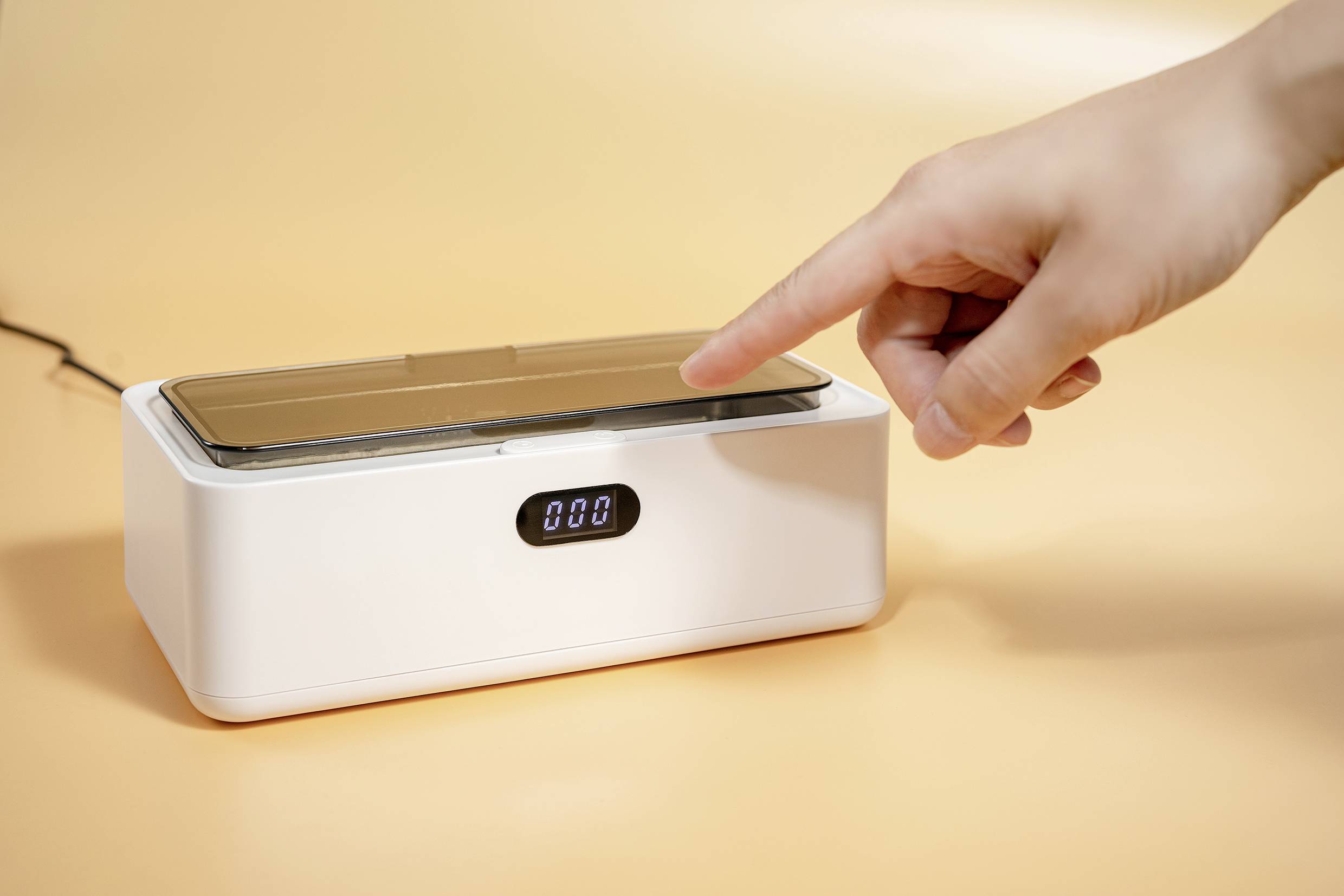 A hand is pointing to a white, rectangular device with a digital display. The device is sitting on a yellow background.