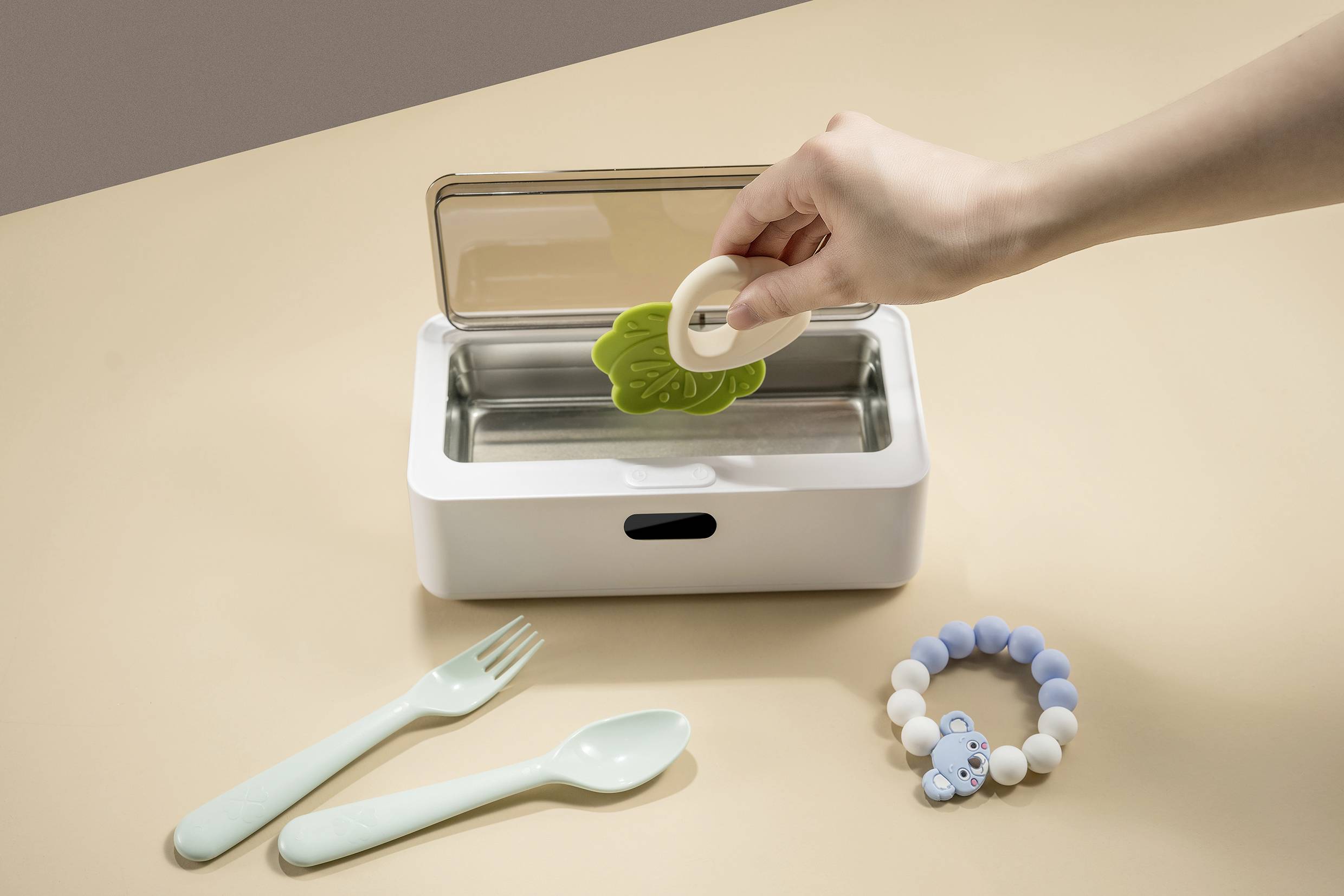 A hand places a green leaf-shaped and a white round teething ring into a white steriliser. Beside them are a spoon, a fork, and a blue-white wristband.