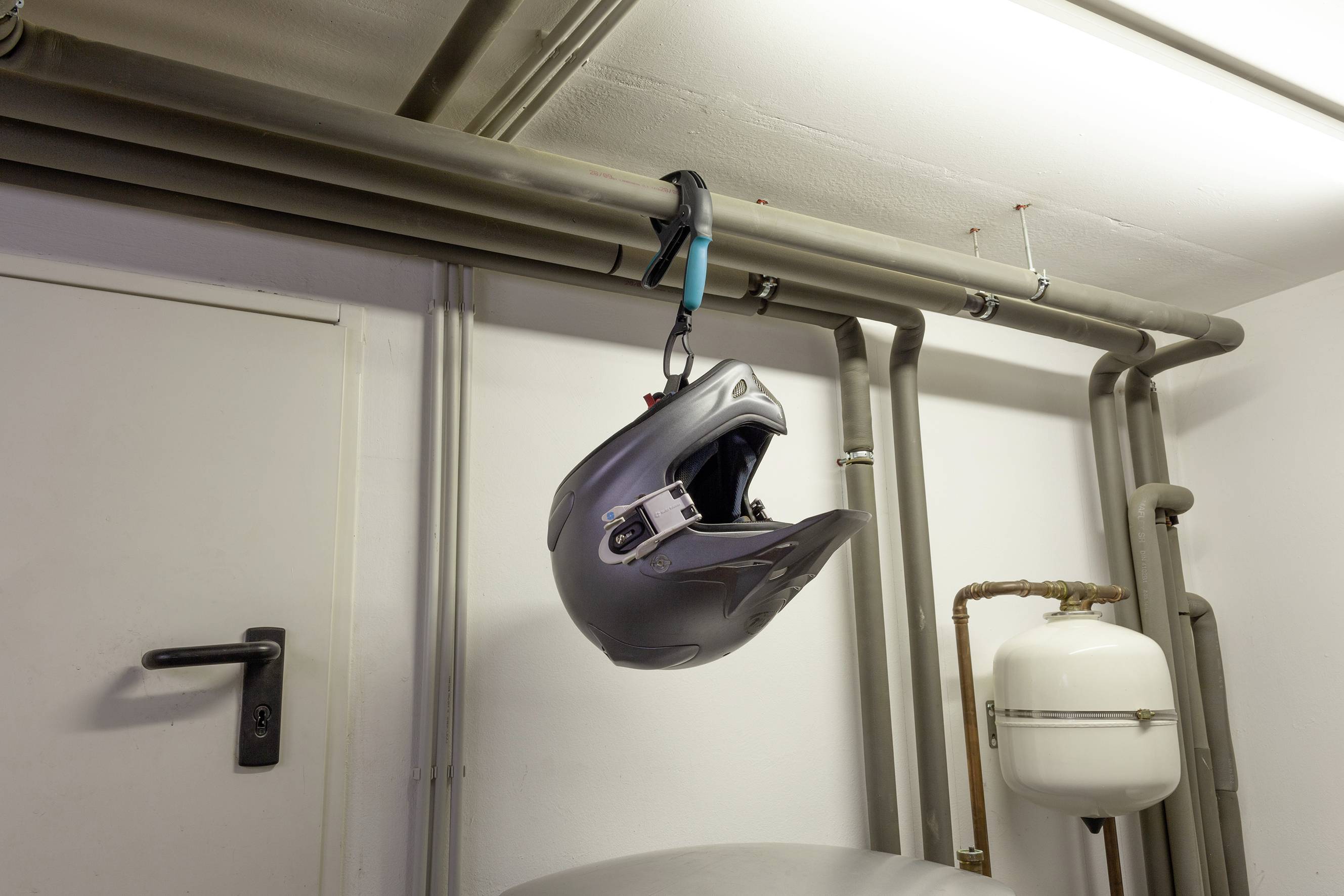 A motorcycle helmet hangs from a pipe in an interior space. The helmet is dark grey and secured with a click fastener. A door is located to the left.