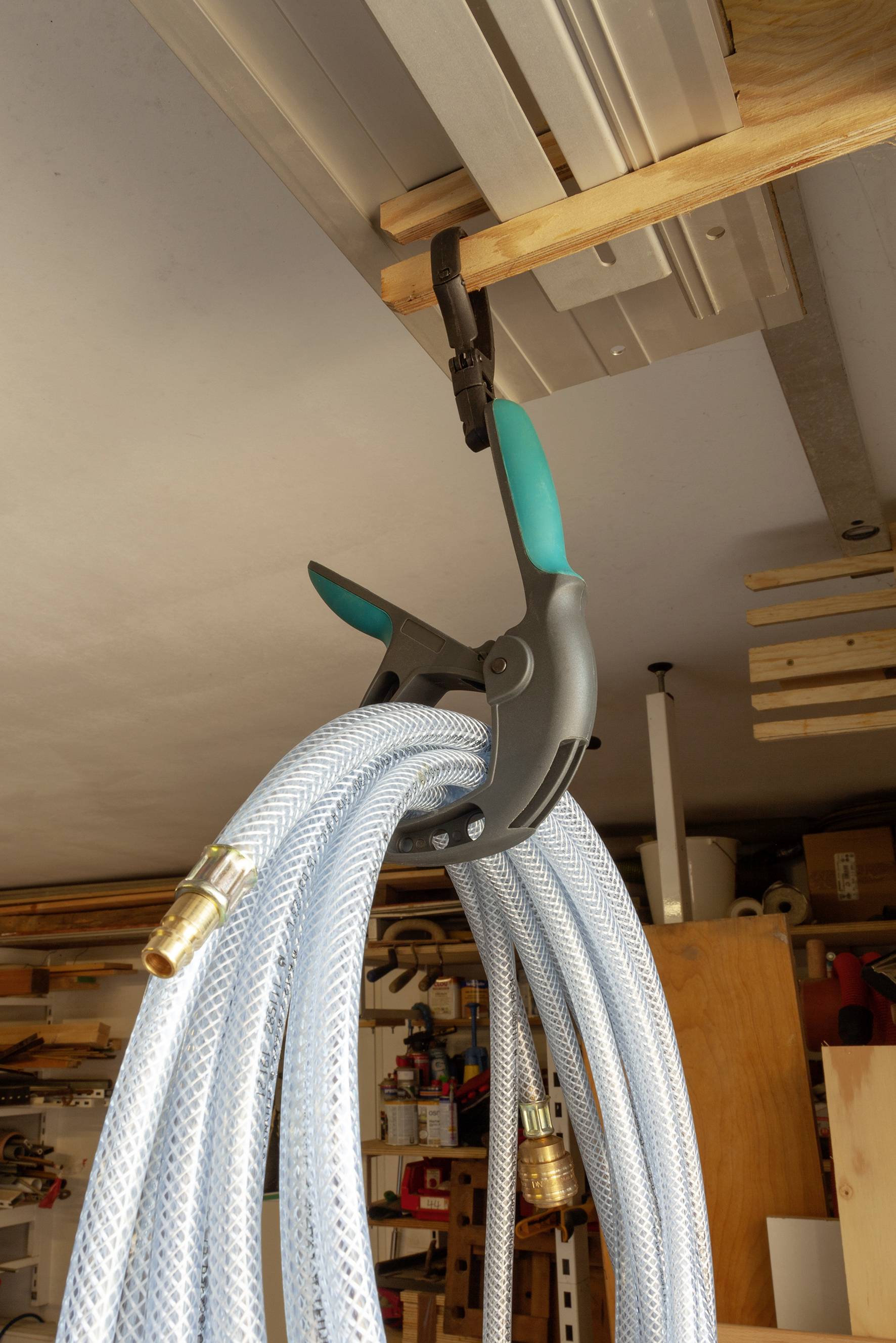 A grey hose is suspended from a wooden shelf with a large clamp in a workshop. Tool-laden shelves are visible in the background.