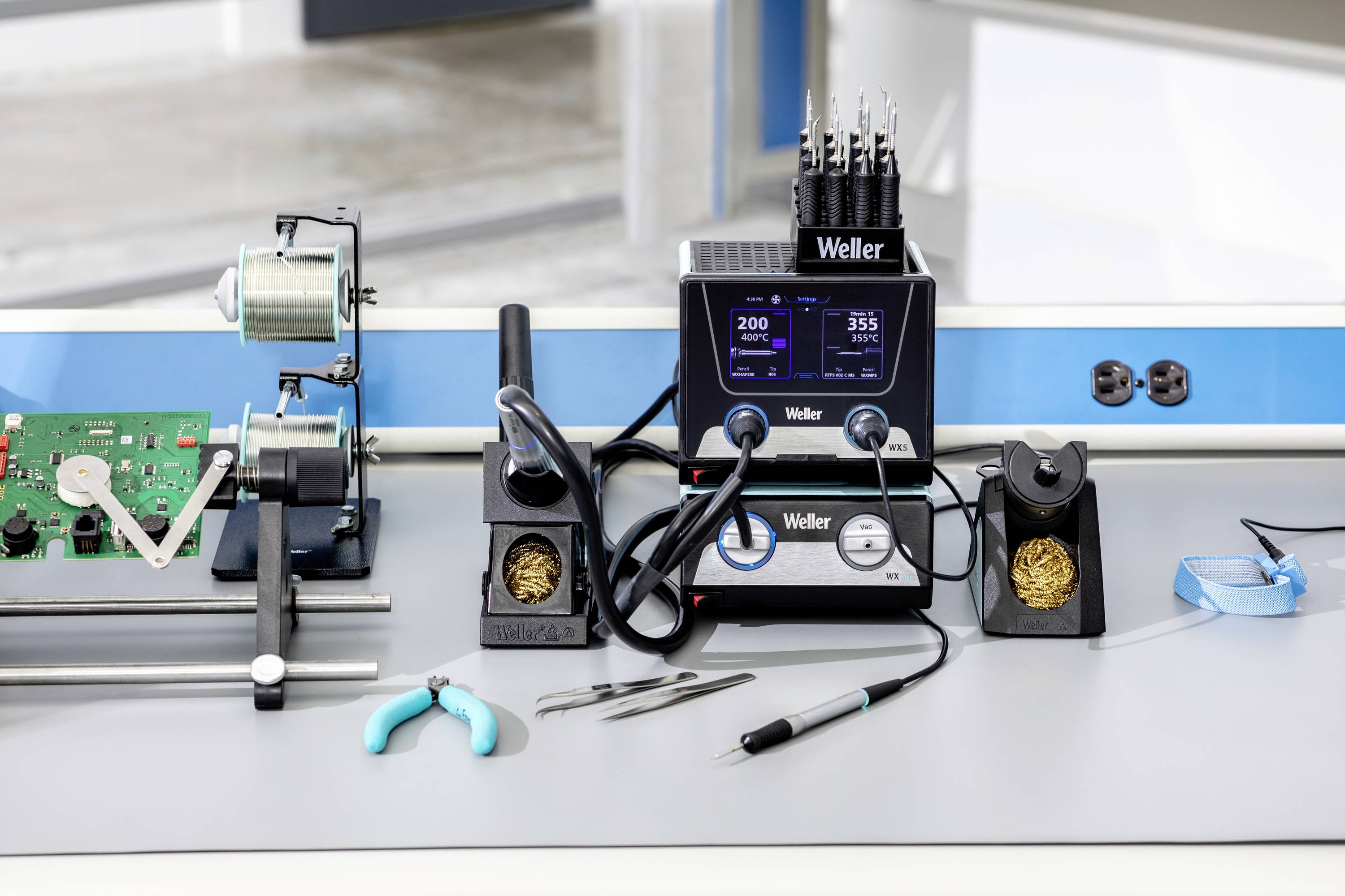Electronic workstation with soldering iron, circuit board in holder, tweezers and pliers. Main device shows temperature display.