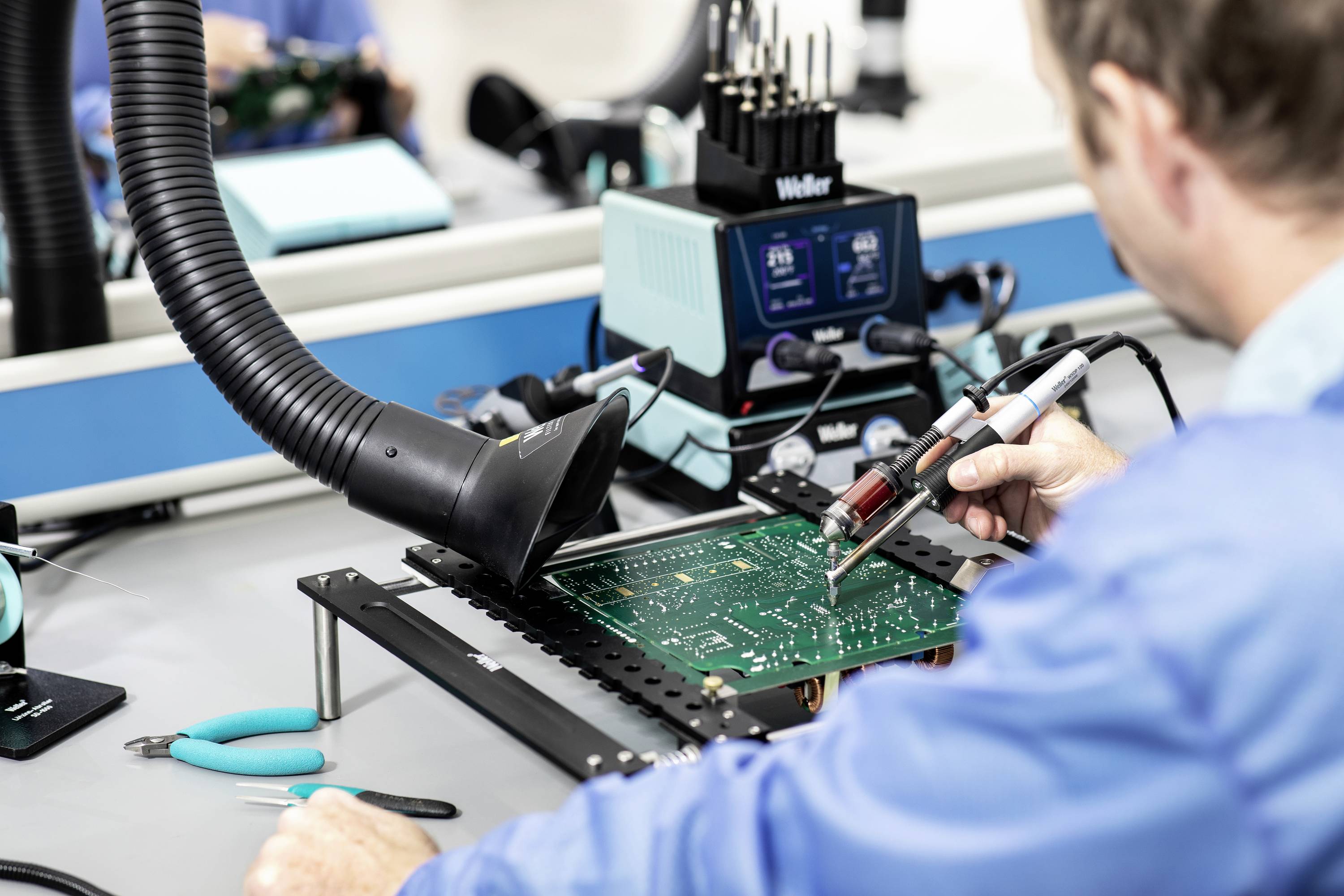A technician solders components onto a circuit board in an electronics workshop. Tools and equipment are visible in the background.