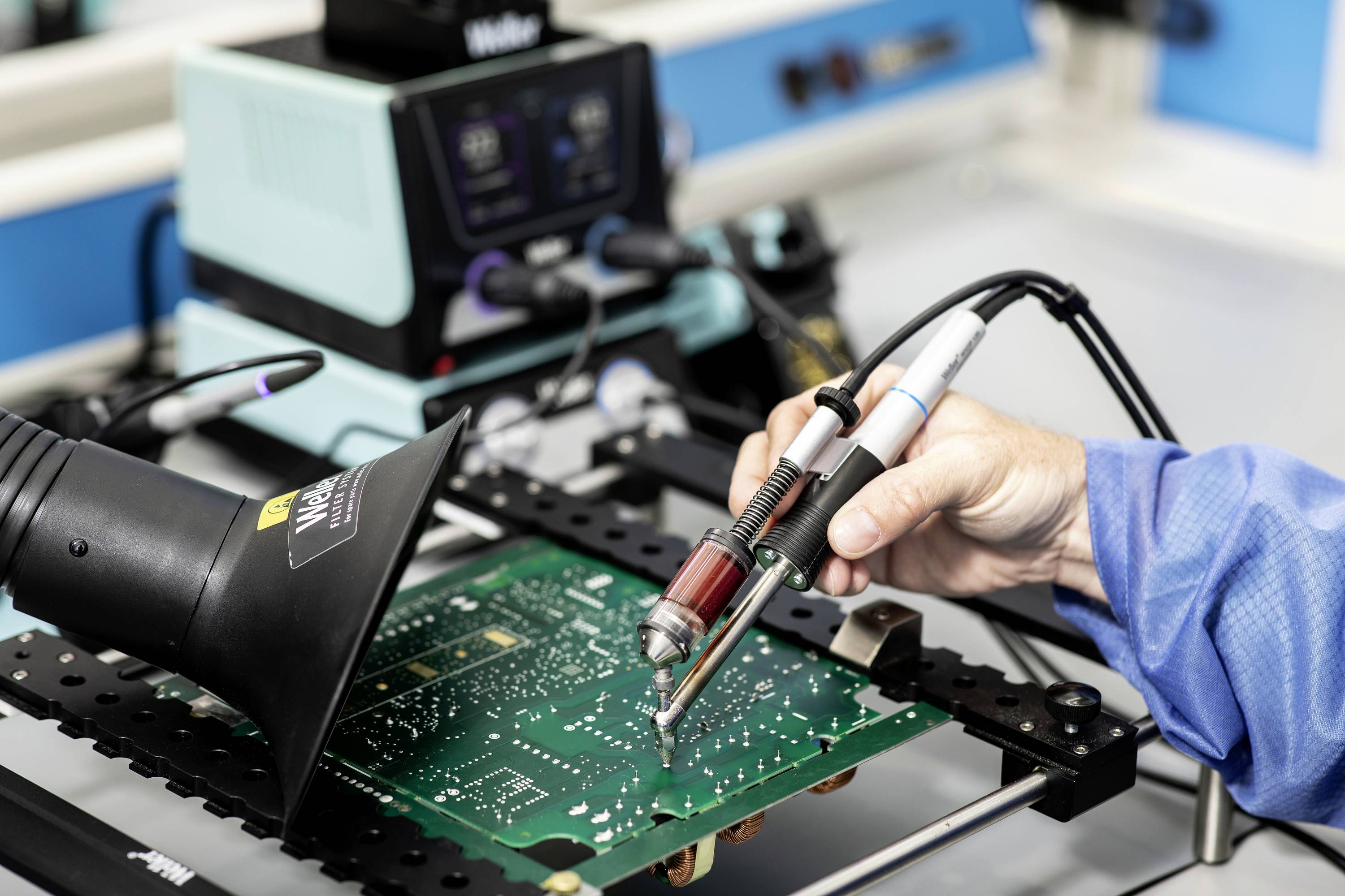 A person is soldering with a soldering iron on a green circuit board in a workshop. A soldering station is visible in the background.