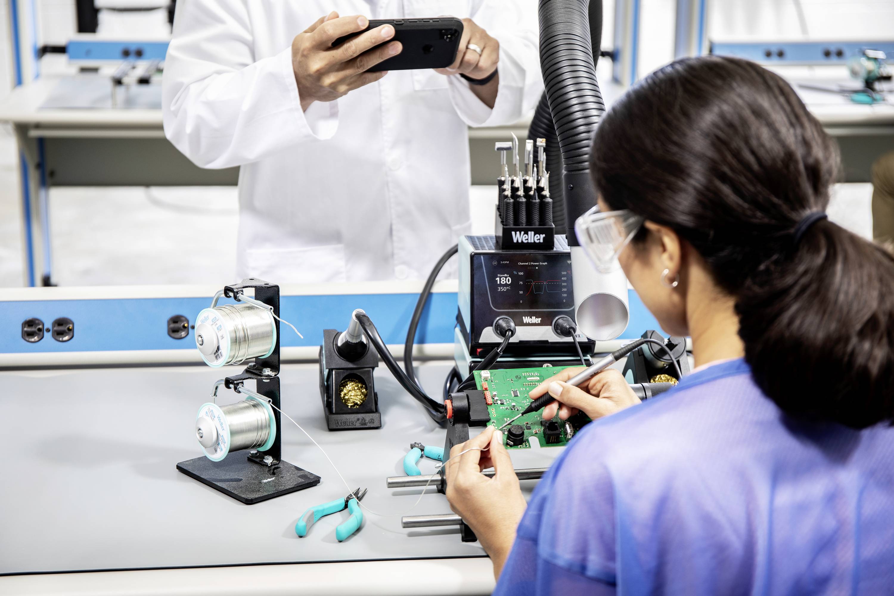 A woman is soldering a circuit board in a workshop. A man in a white lab coat is photographing her with a smartphone.