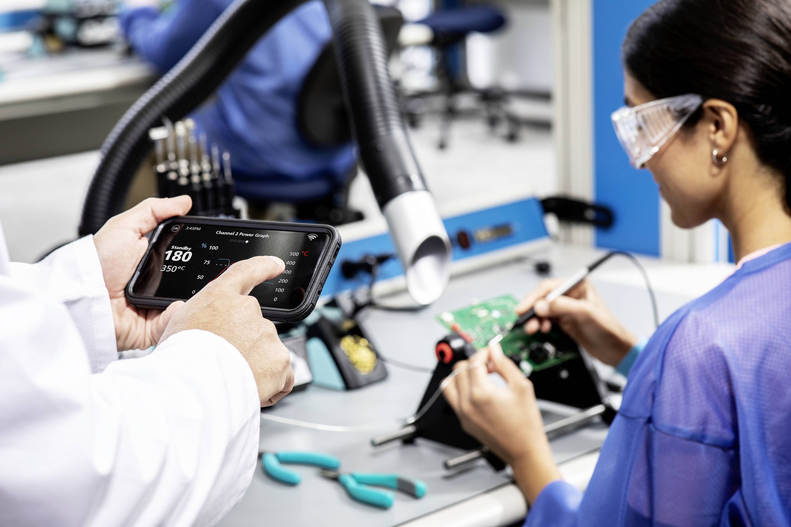 A person is soldering a circuit board; someone nearby is using a tablet to display ventilation settings. Work environment with tools.
