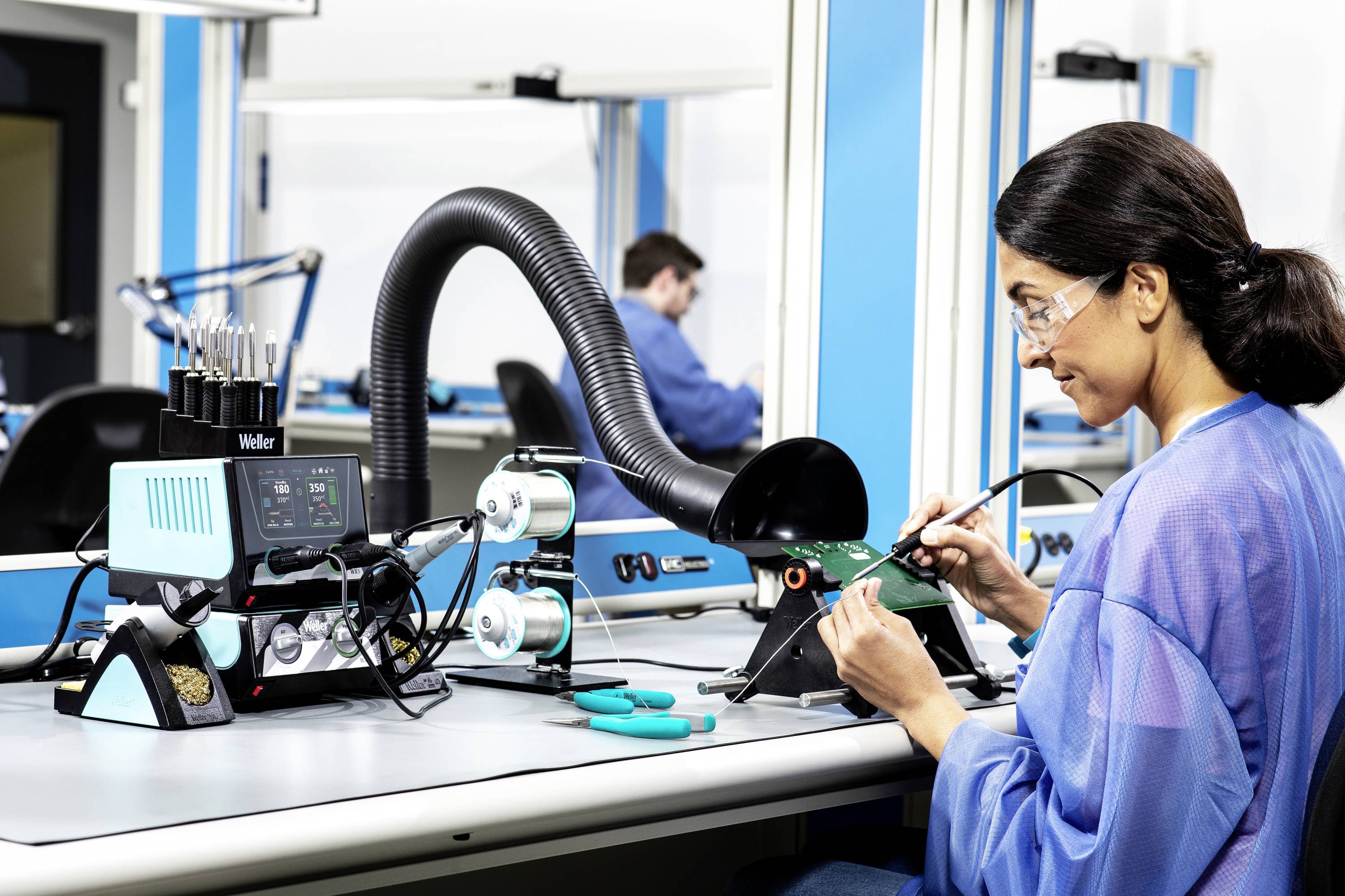 A woman wearing safety glasses is soldering a circuit board in an electronics laboratory. Another person can be seen in the background.