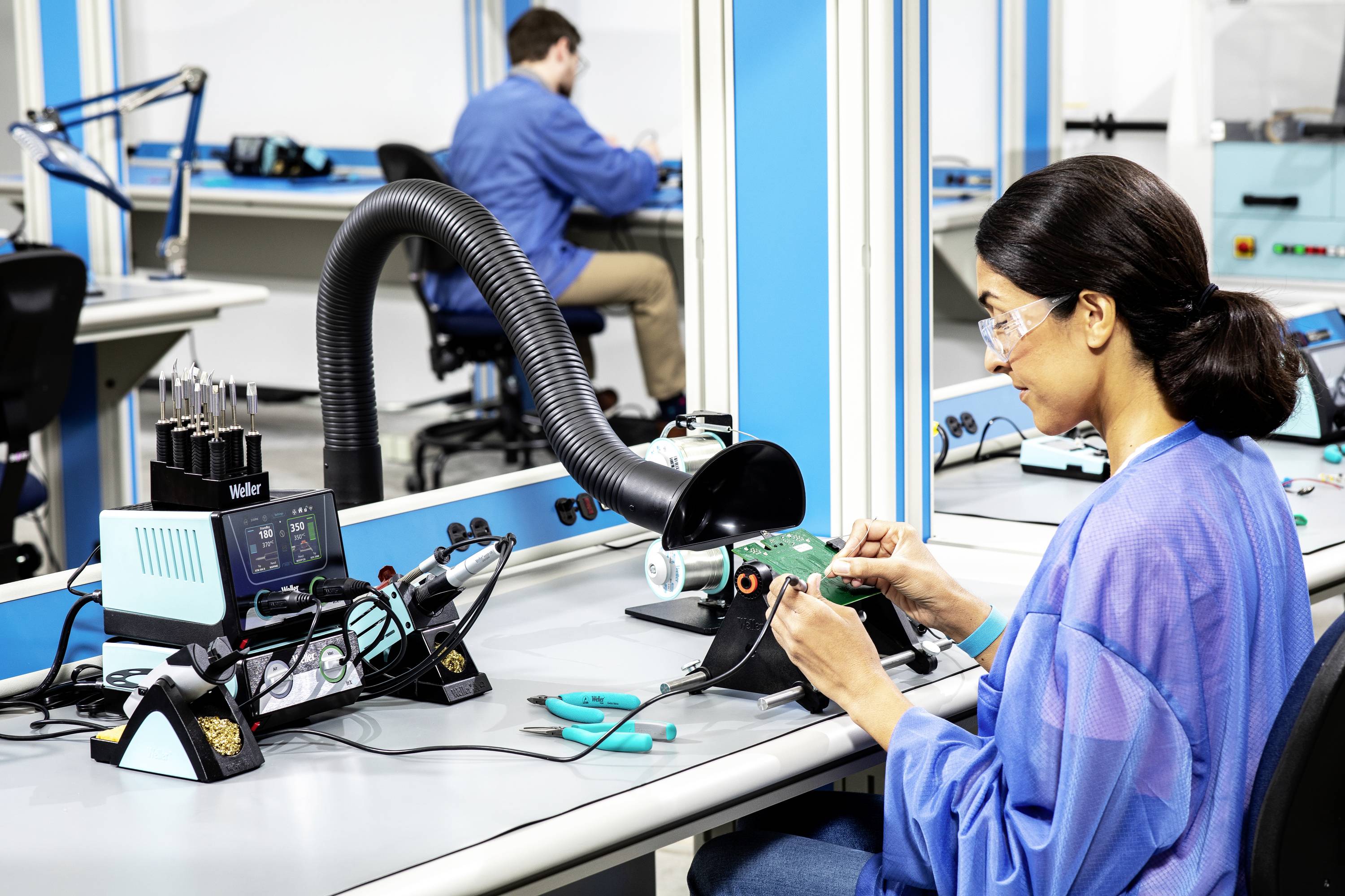 A woman in protective clothing is soldering a circuit board at a workstation. In the background, another person is working at a table.