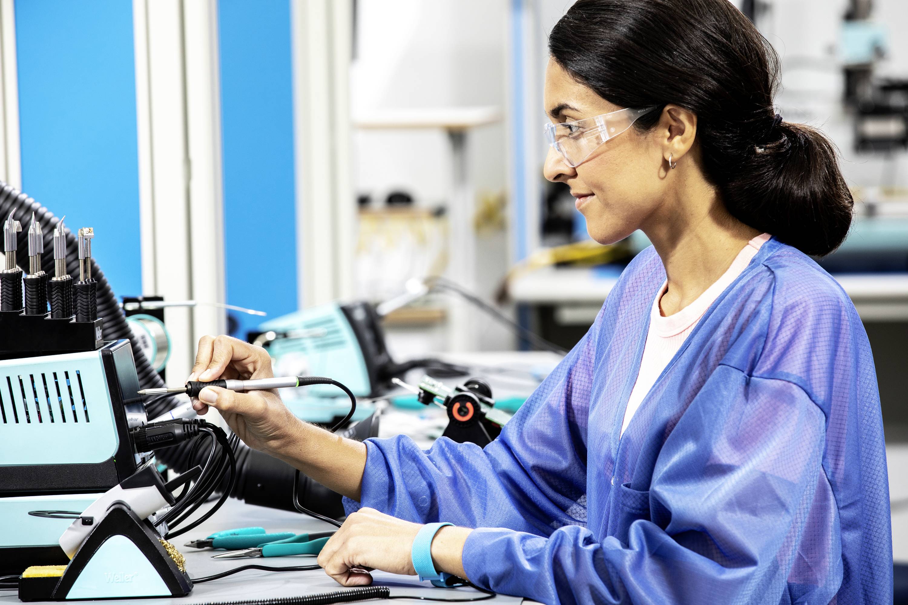 A woman wearing safety glasses and blue workwear is working with a soldering iron on electronic components in a laboratory.