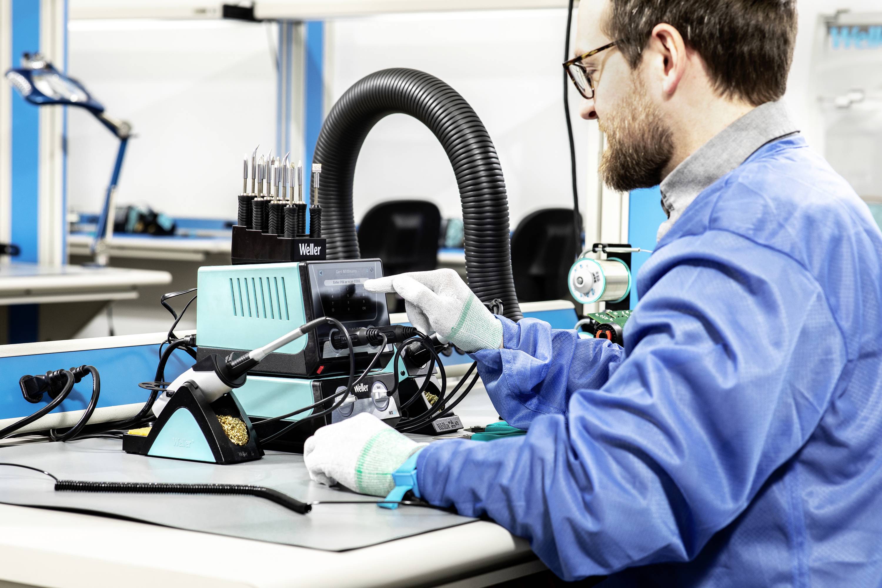 A person wearing blue workwear is sitting at a table and operating a soldering device in a laboratory environment.