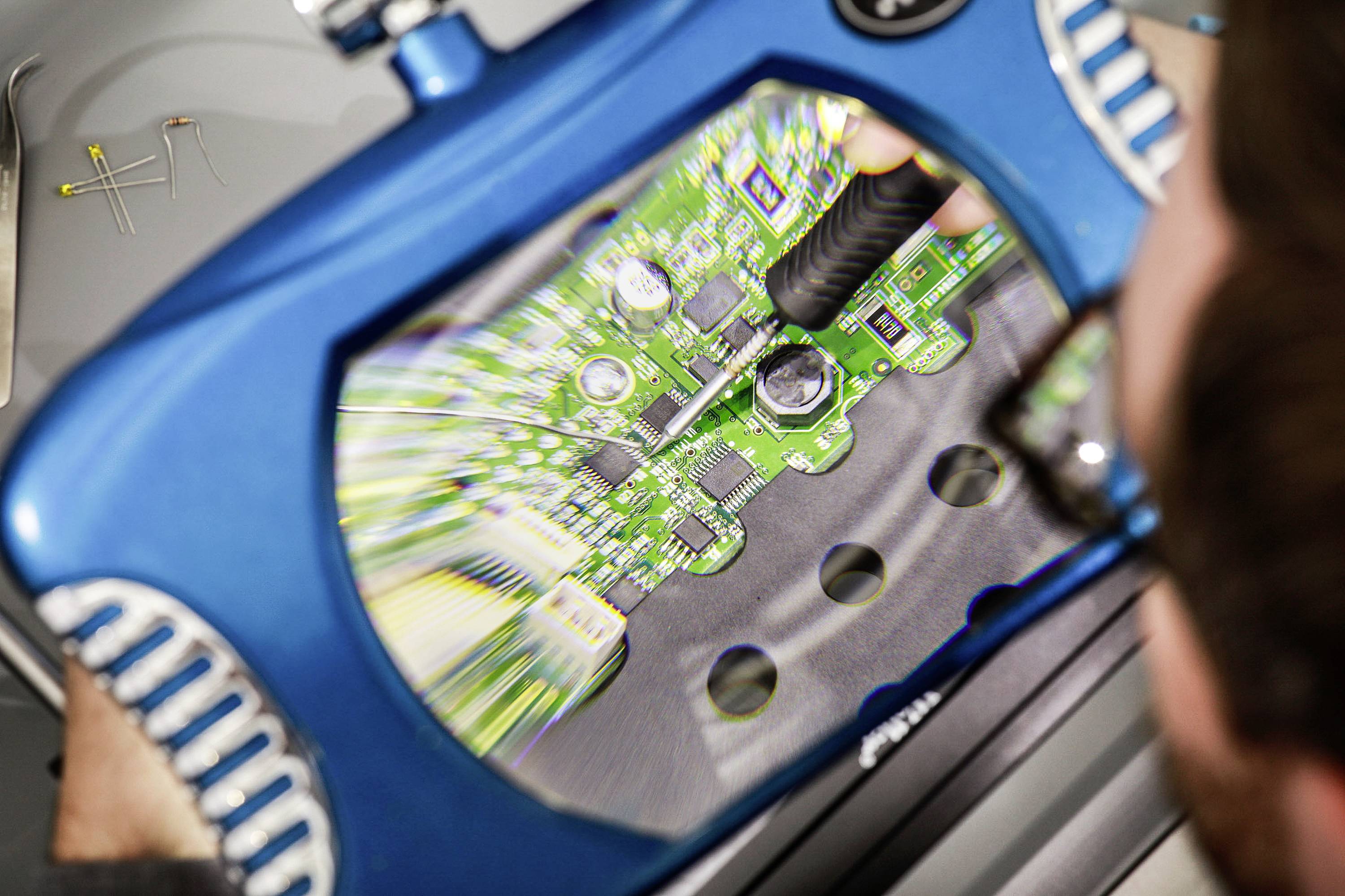 A person is examining a green circuit board through a blue magnifying glass. The board displays complex circuit components.