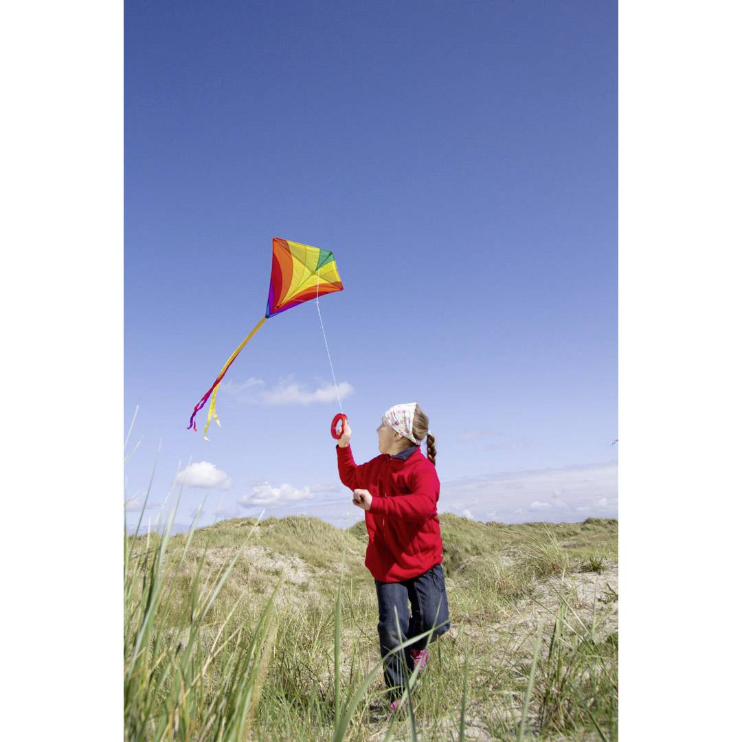 A girl in a red jumper flies a colourful kite in a grassy sand dune area on a sunny day.