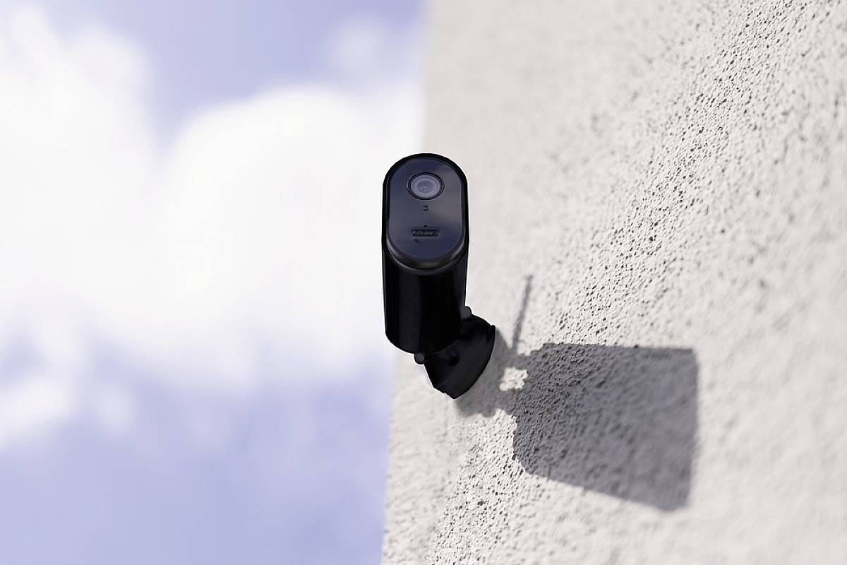 A black surveillance camera is mounted on a grey wall and pointing towards the sky. The sky is blue with some white clouds.