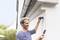 A man is mounting a camera on a house wall and looking at his mobile phone. The surroundings are green and the sky is clear.