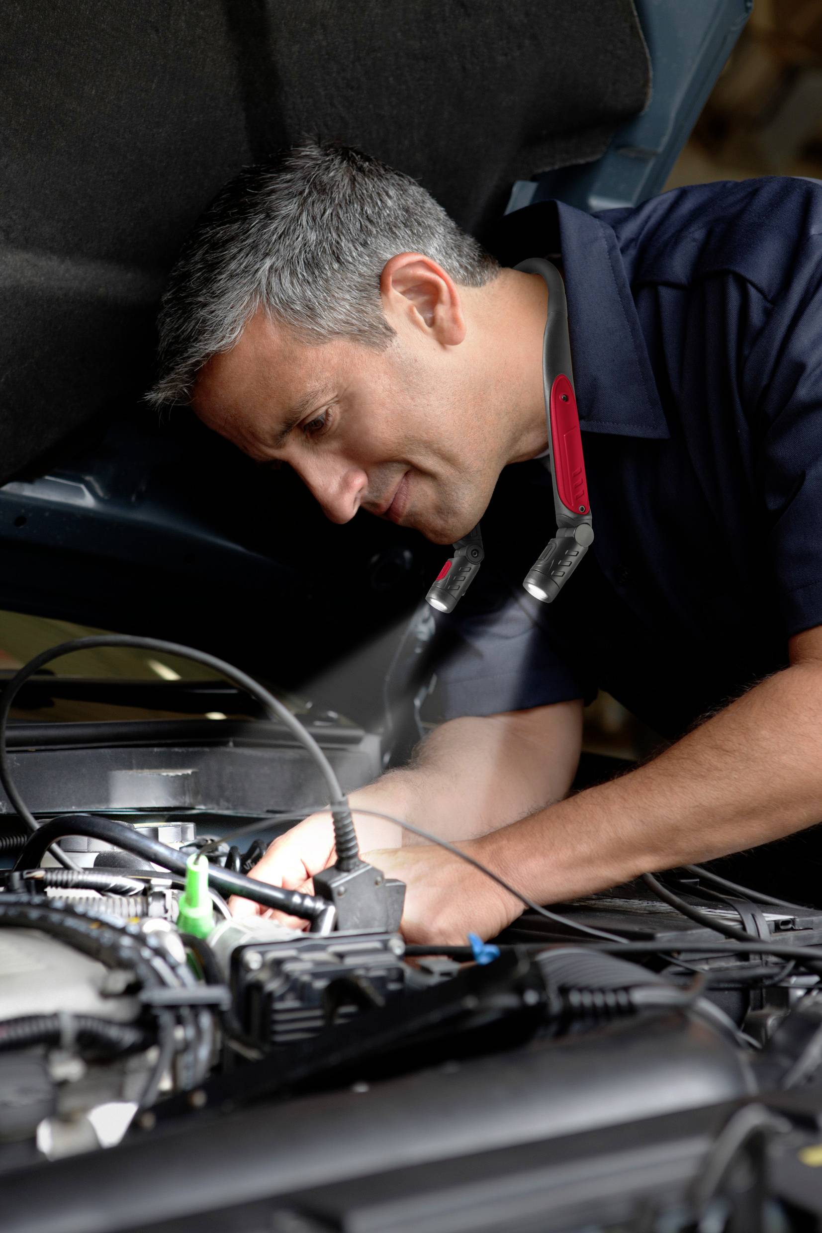 A mechanic is repairing a car's engine. He is wearing a head torch around his neck that illuminates the work area.