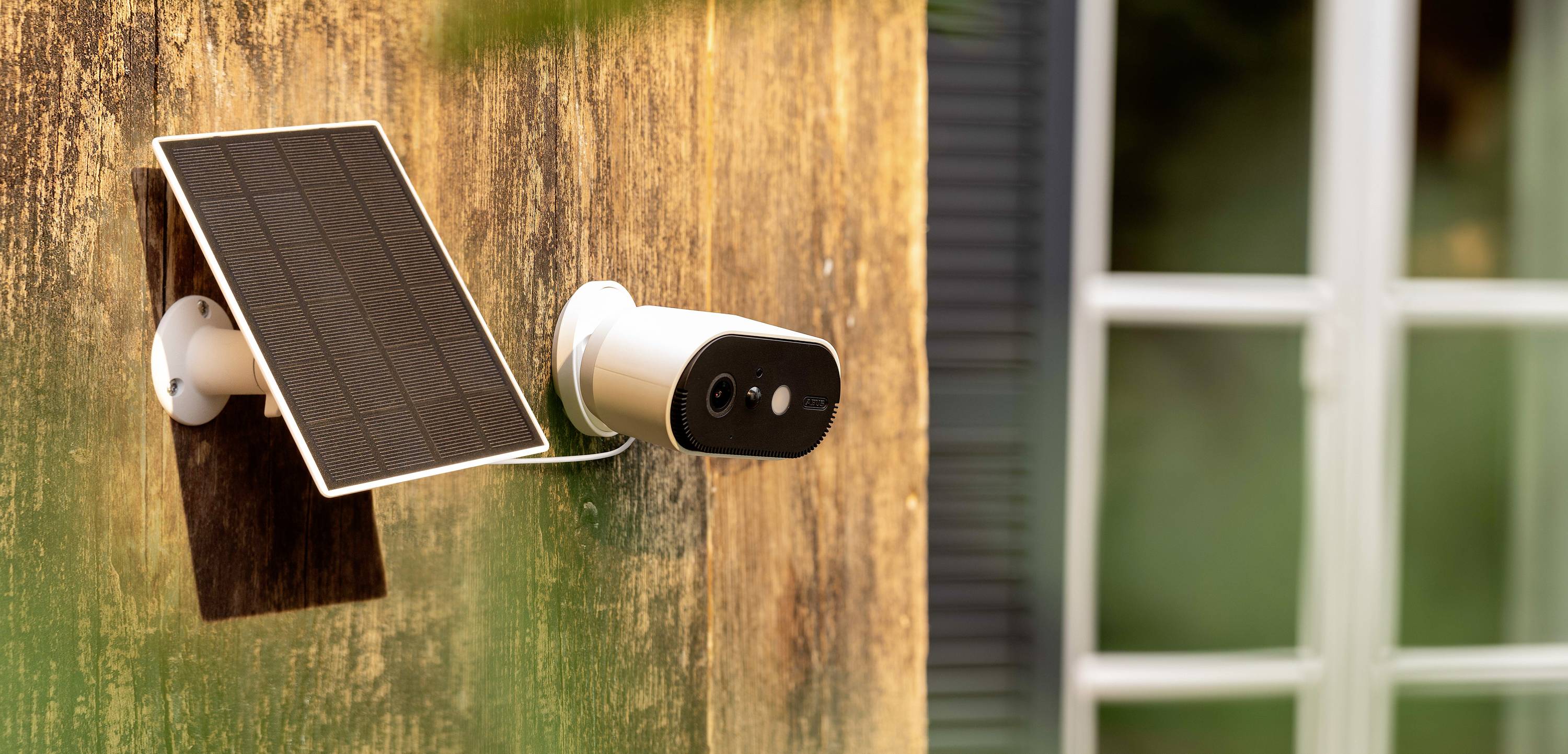 A security camera with a solar panel is mounted on a wooden house wall, with a window in the background.