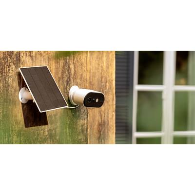 A security camera with a solar panel is mounted on a wooden house wall, with a window in the background.