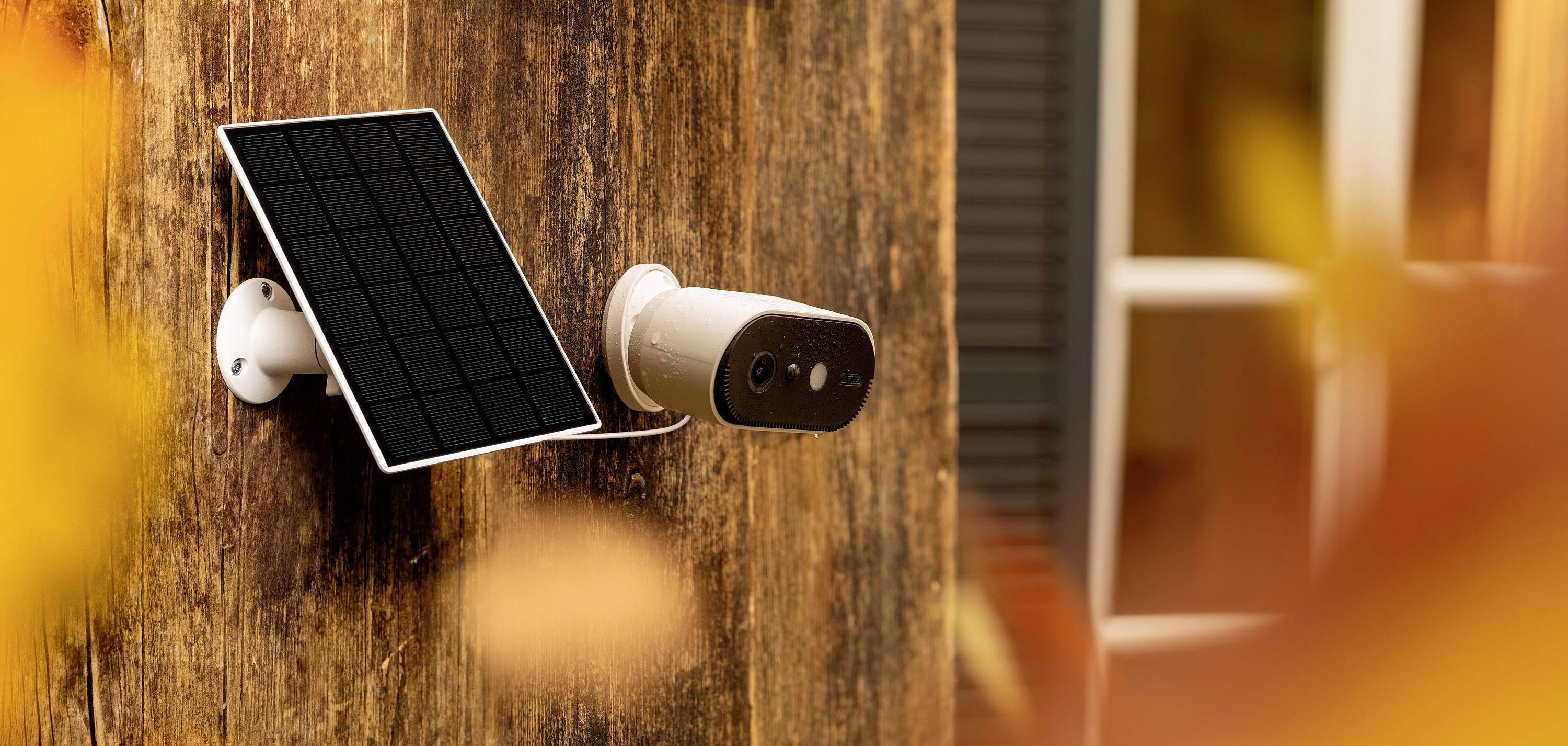 A white security camera with a solar panel is mounted on a wooden facade. Blurred autumn leaves are visible in the foreground.