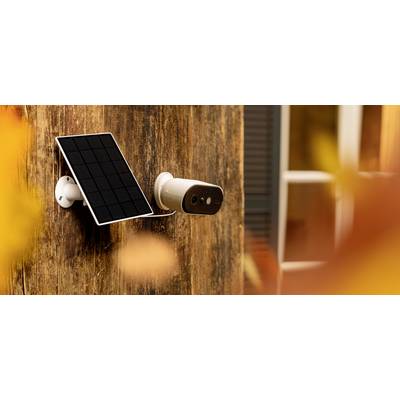 A white security camera with a solar panel is mounted on a wooden facade. Blurred autumn leaves are visible in the foreground.