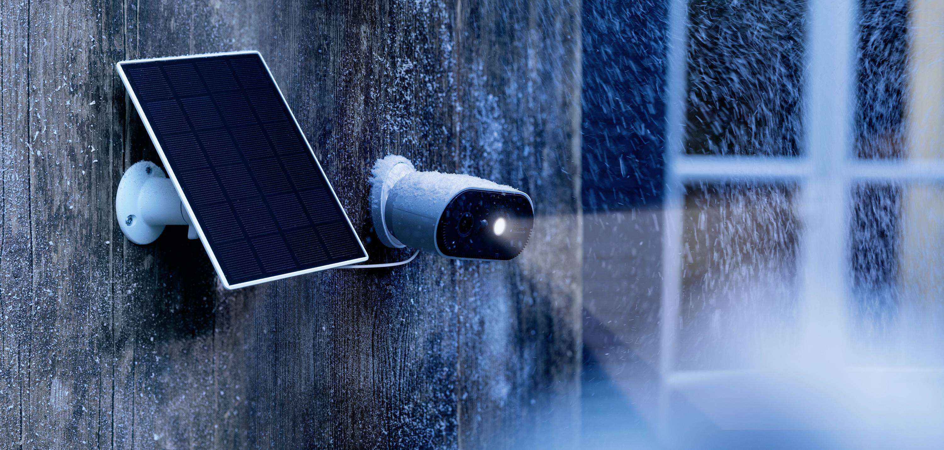 Security camera with solar panel, installed on a house wall during snowfall, illuminating an area in the foreground.