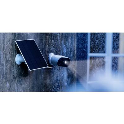 Security camera with solar panel, installed on a house wall during snowfall, illuminating an area in the foreground.