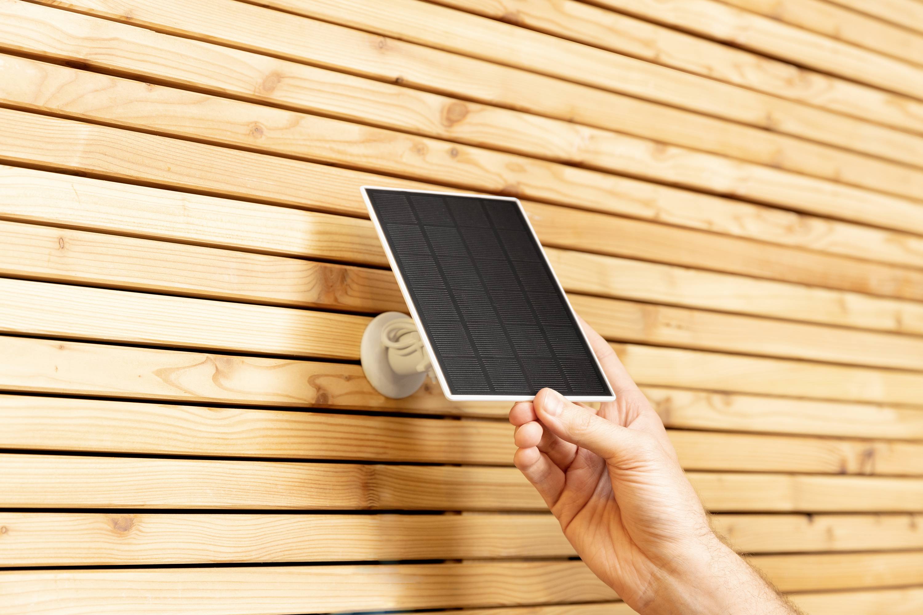 A hand holds a small solar panel in front of a wooden lattice wall.