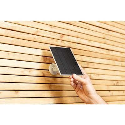 A hand holds a small solar panel in front of a wooden lattice wall.