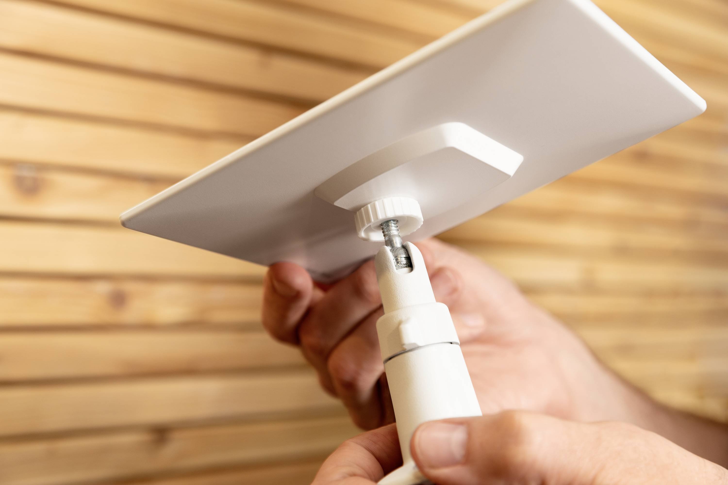 A person is holding and adjusting a rectangular, white satellite dish in front of a wooden wall.