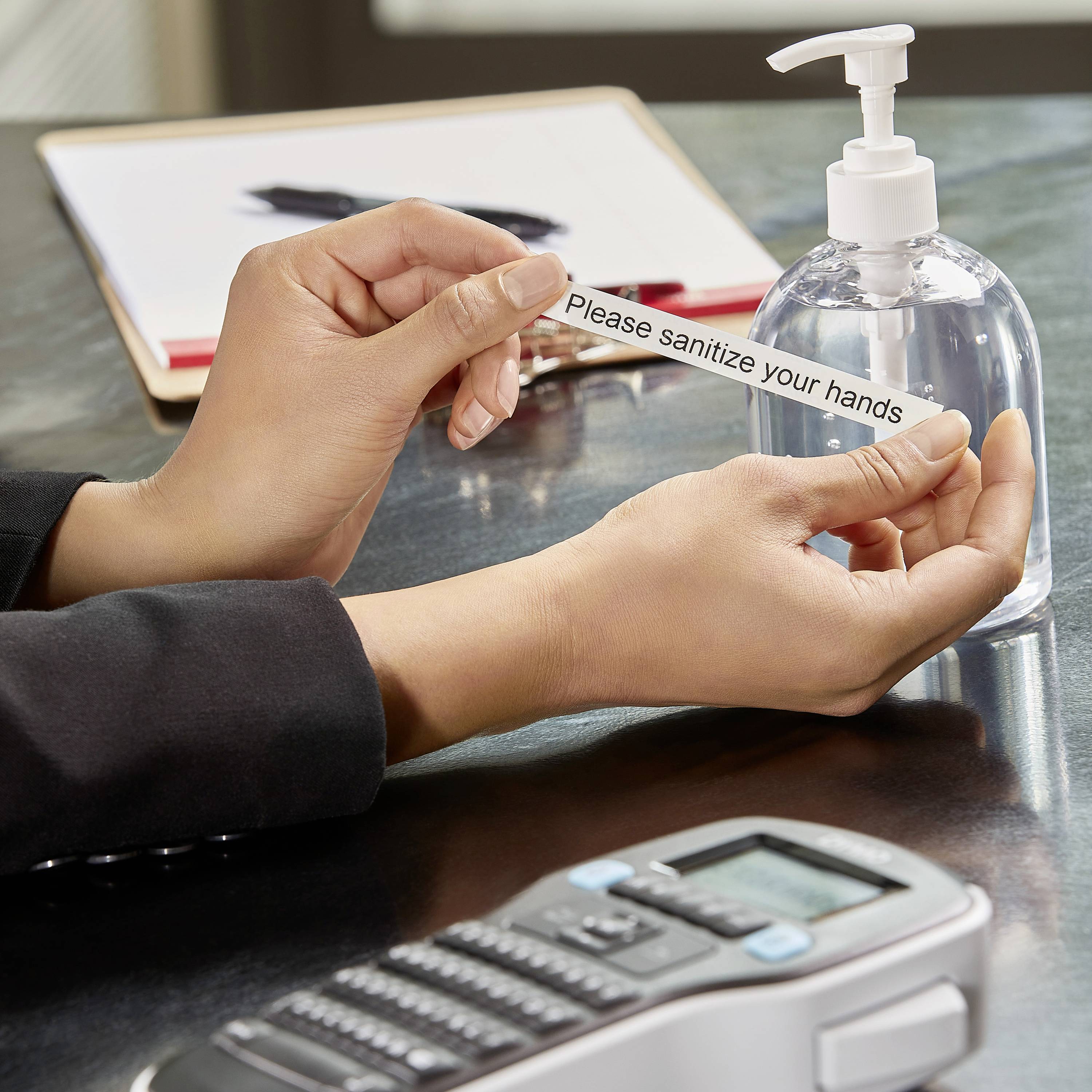 Close-up of hands holding a note reading 'Please sanitize your hands' in front of a hand sanitiser dispenser.