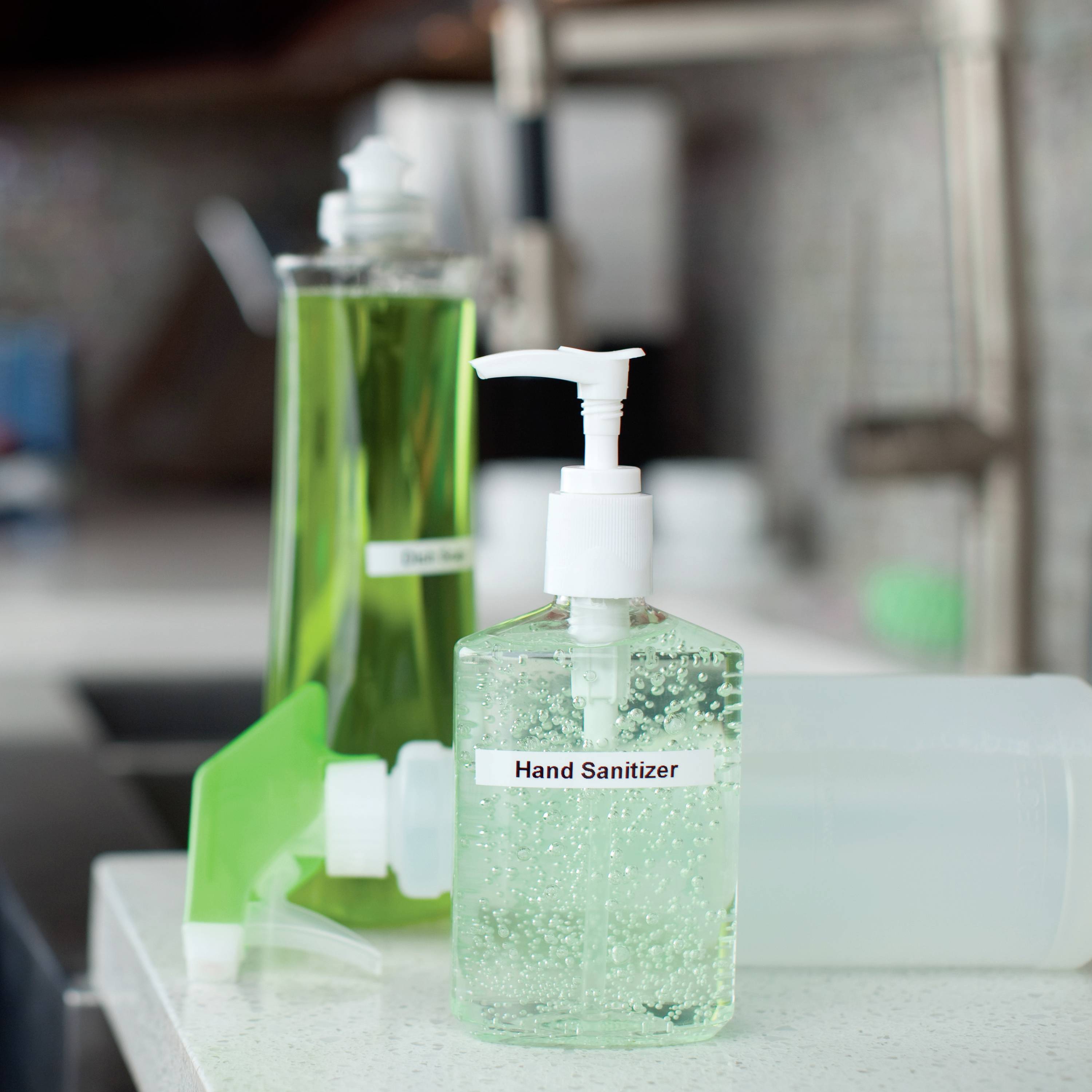 In the foreground stands a transparent dispenser with 'Hand Sanitiser', filled with a clear liquid, in a kitchen.