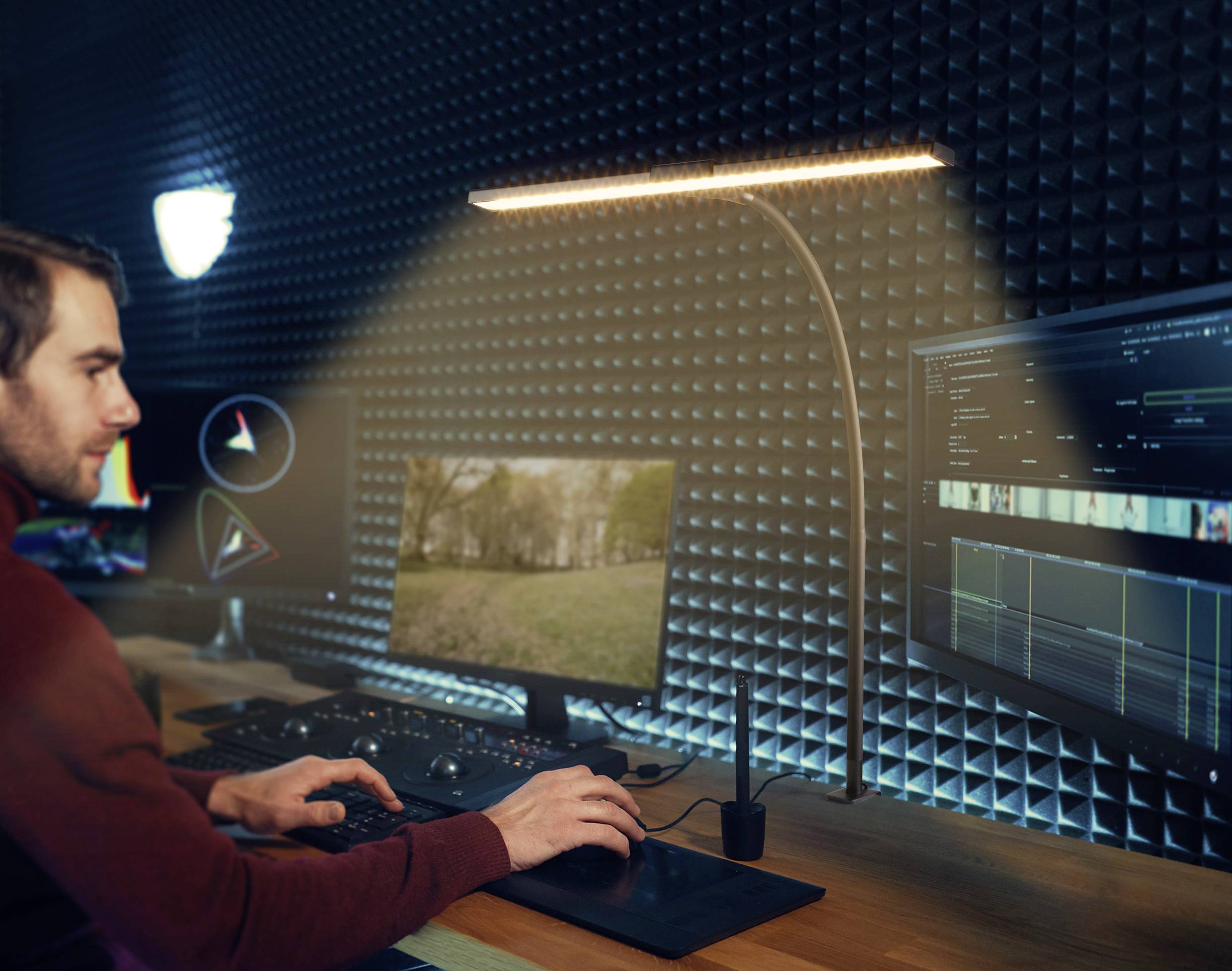 A man sits at a computer workstation in a dimly lit room with multiple monitors. He is working intently at his desk.
