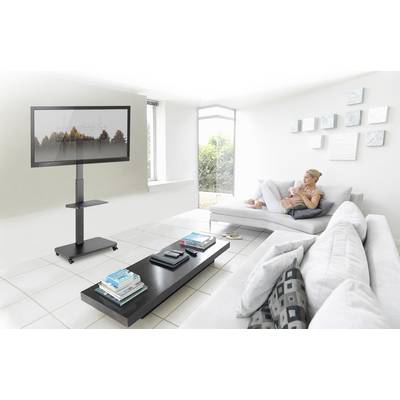 A woman is sitting on a sofa in a bright living room, drinking coffee and watching a television that is on a stand.