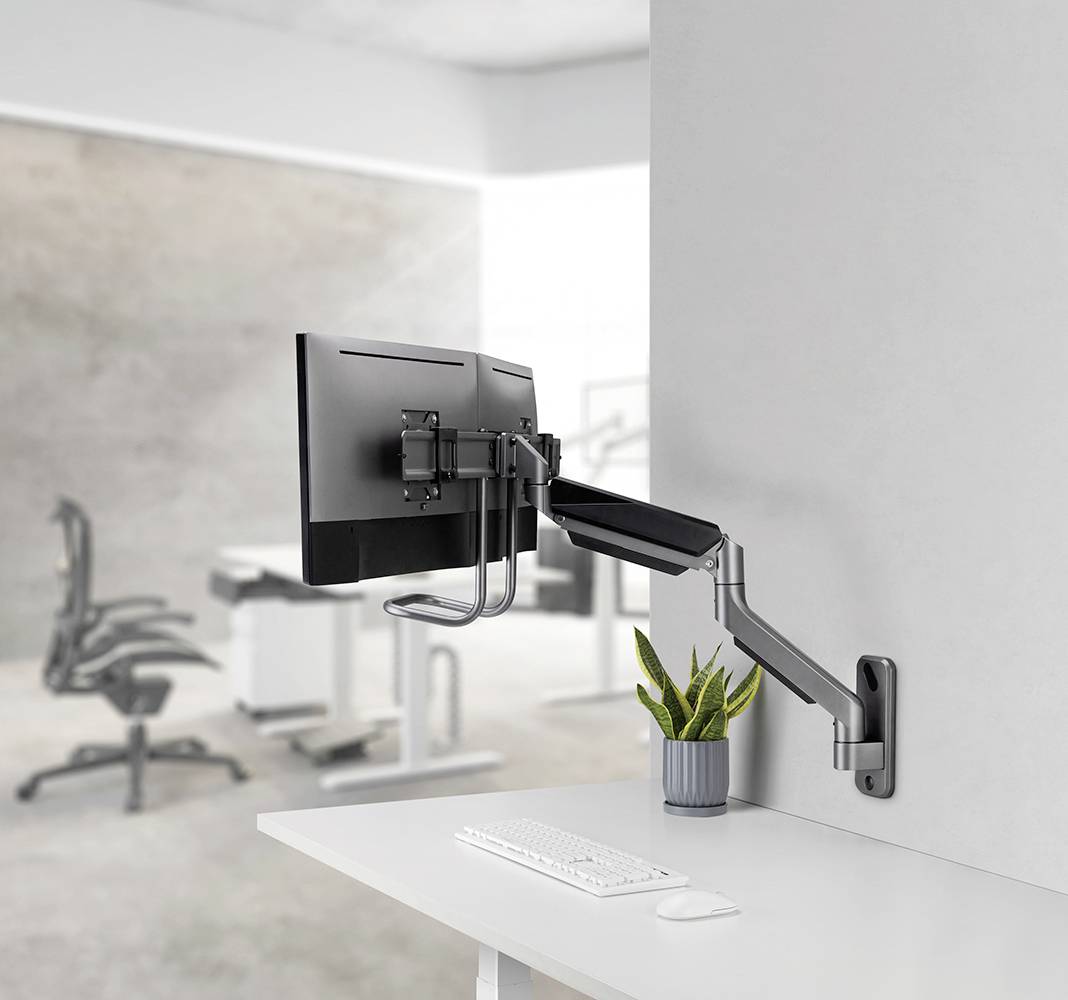 A computer monitor is mounted on an adjustable wall arm. Underneath are a keyboard and a small plant.