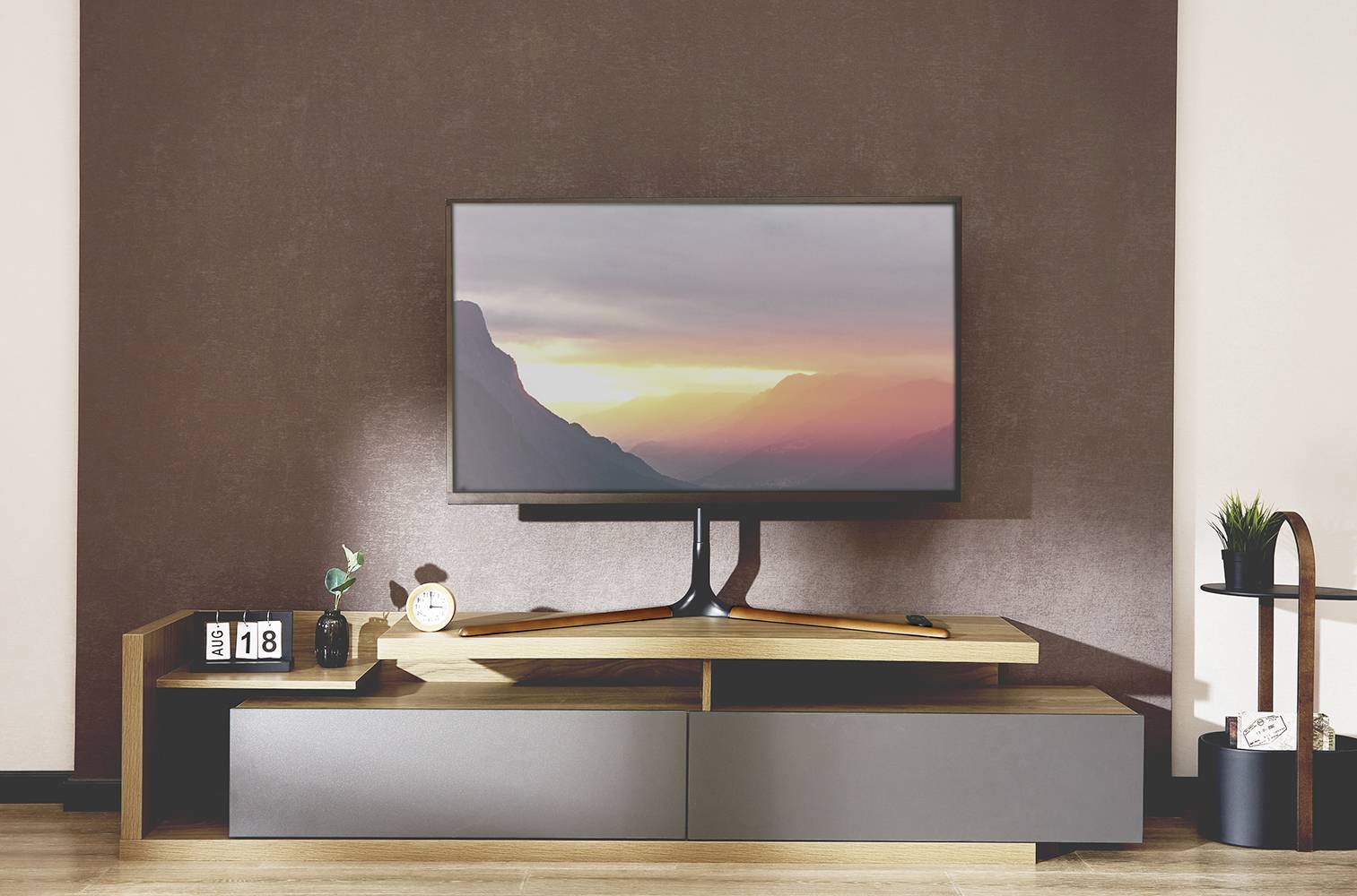 Television on a modern TV stand, displaying a mountain landscape at sunset. Decorative elements such as a clock and plant beside it.
