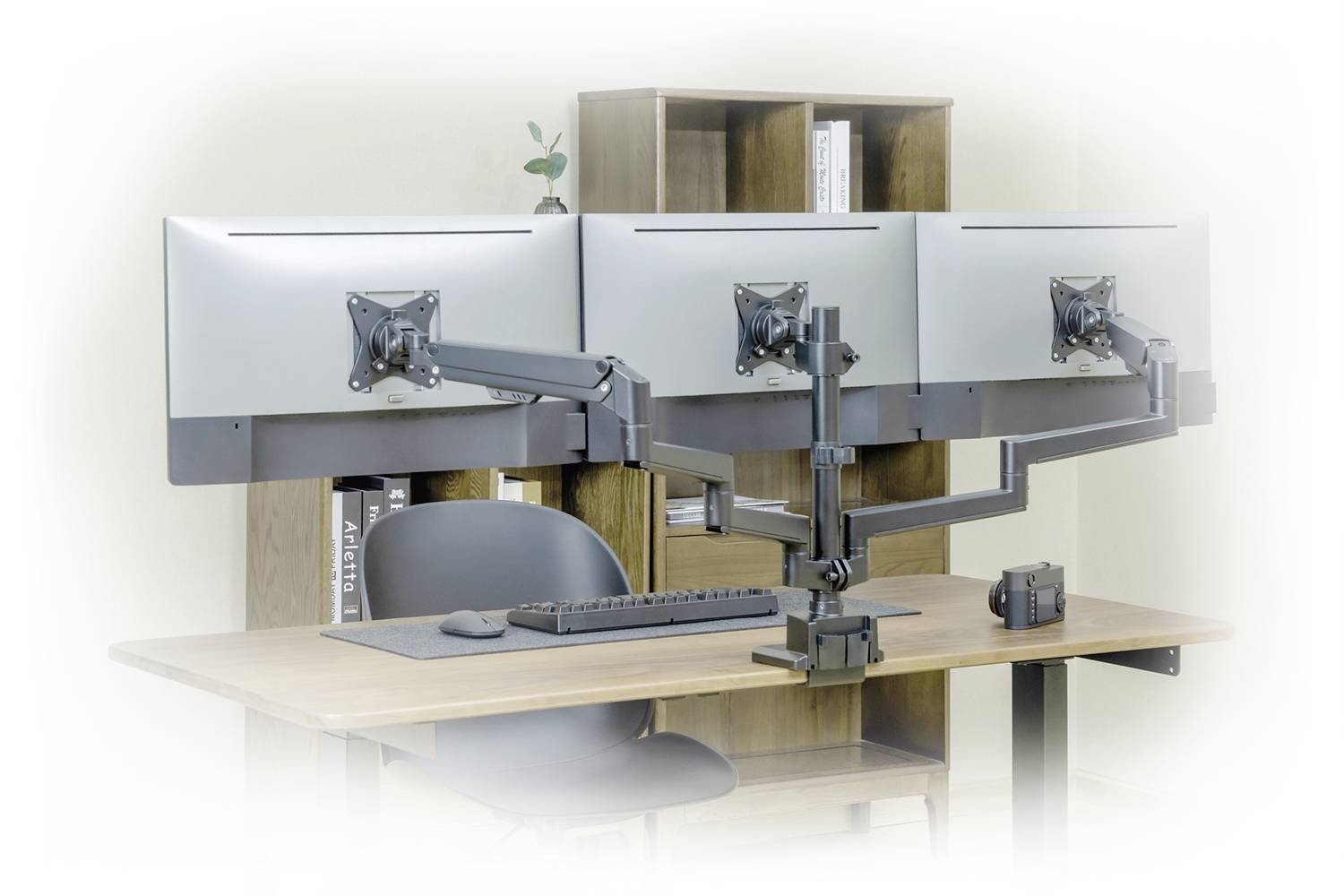 Three monitors are mounted on an adjustable stand above a desk. Beneath them are a keyboard and a camera.