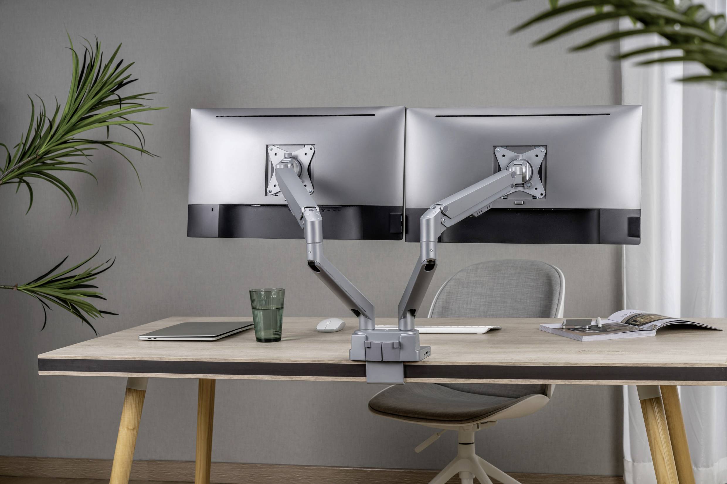 A modern desk with two large monitors on adjustable mounts, a laptop, a glass, and a magazine.