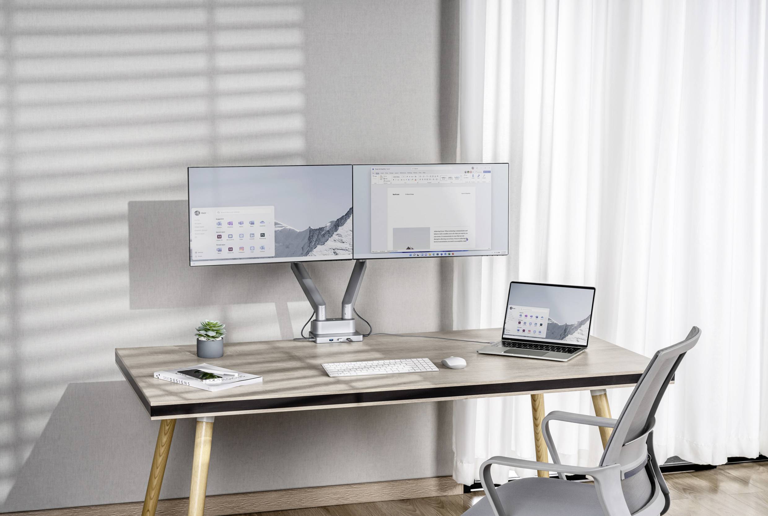 A desk with two monitors, a laptop, keyboard and mouse. In the background, large windows with white curtains.