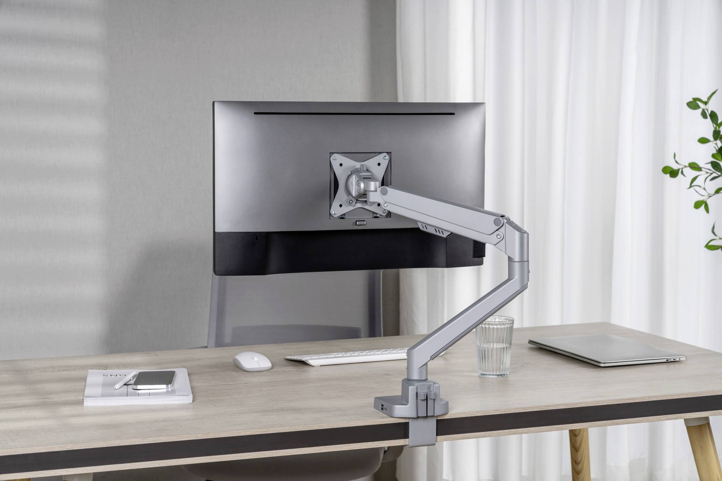 A desk with a monitor on an adjustable arm, a keyboard, a glass of water, a laptop and a book with a smartphone are placed on it.