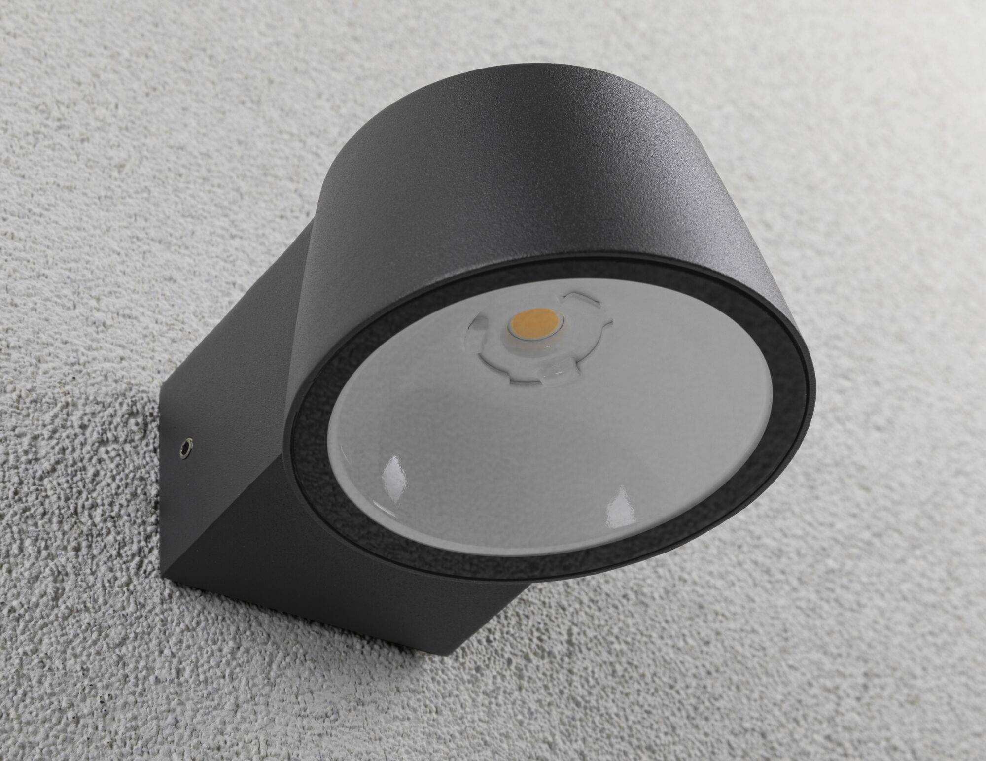 A modern, black outdoor light on a grey wall. It has a round shape and illuminates downwards.
