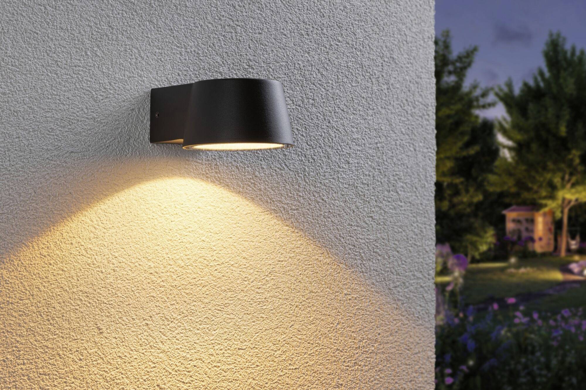 A black wall light illuminates on an external wall, casting light on the area beneath it. In the background, a garden can be seen at dusk.