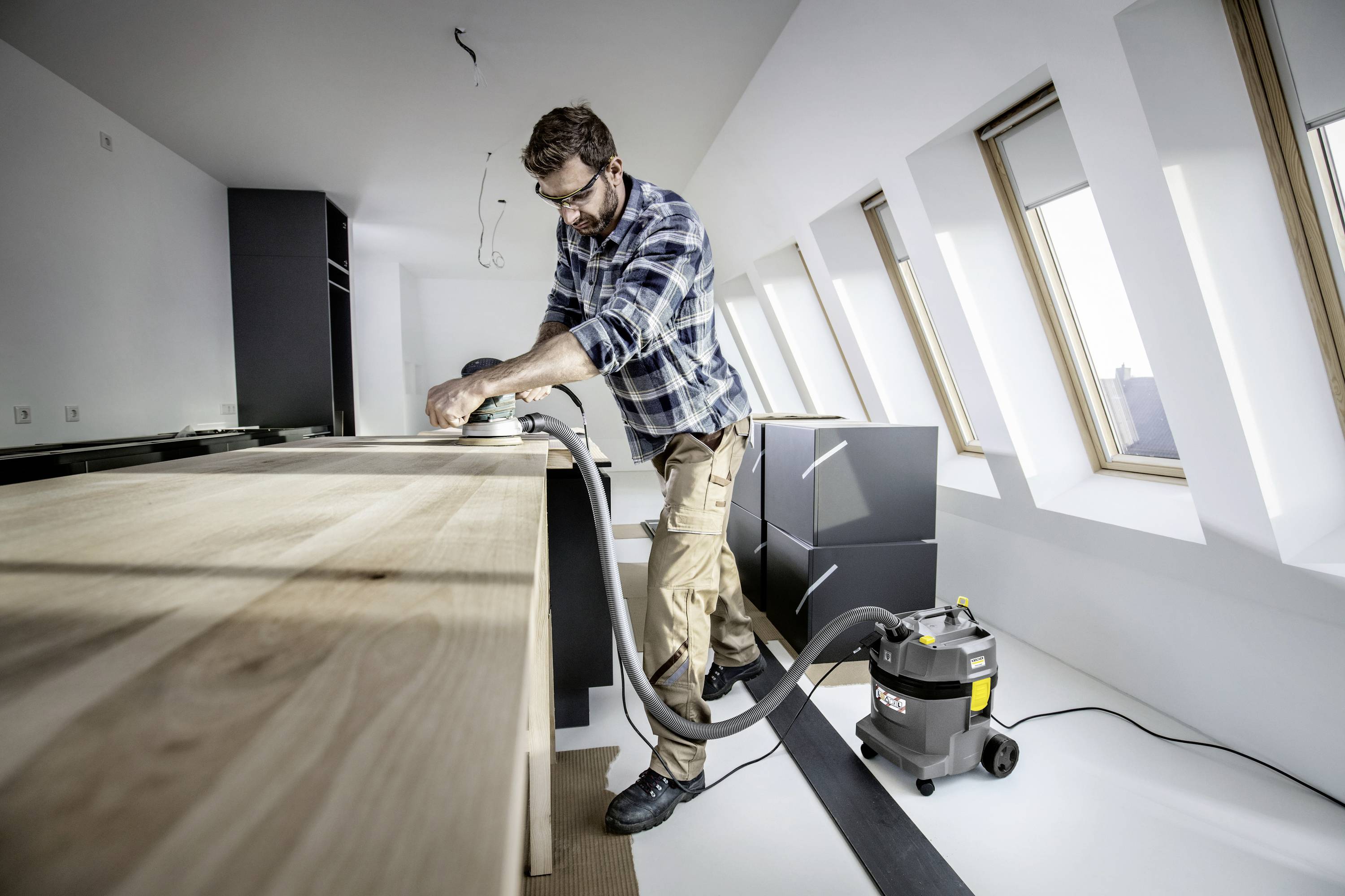 A man is sanding a wooden board in a modern, bright kitchen. A vacuum cleaner is standing next to him to capture the dust.