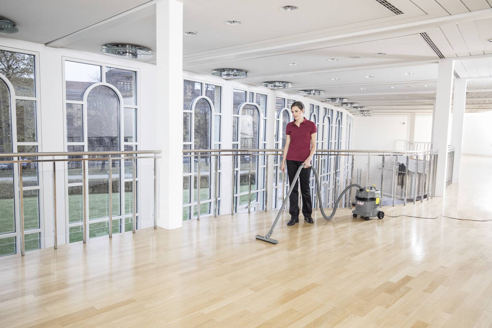 A person in a red shirt is using a vacuum cleaner on a polished wooden floor in a spacious, modern building with large windows.